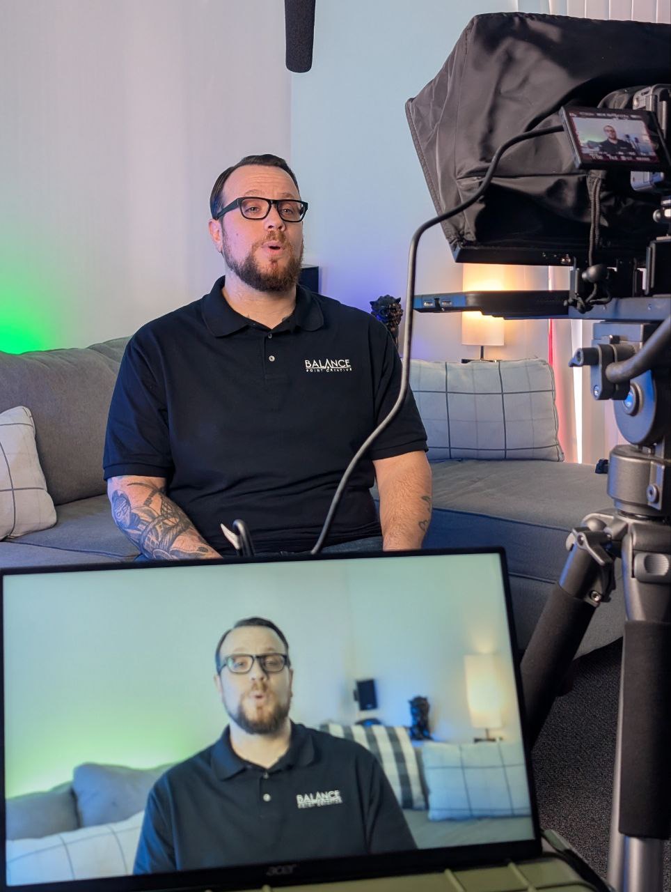 Man with glasses and beard sitting on a couch, being filmed with a camera, with a laptop showing his image.