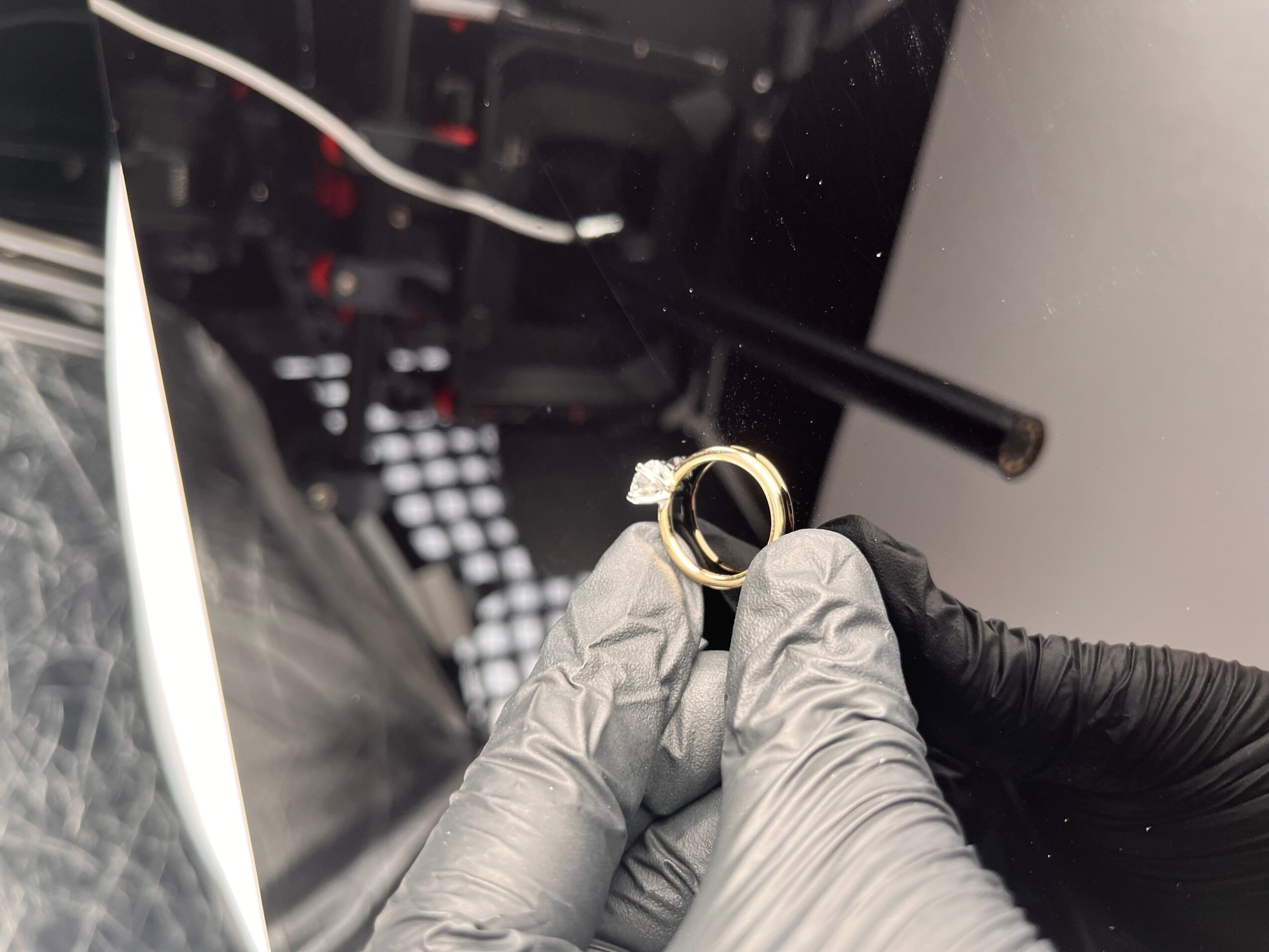 Person wearing black glove holding a small gold ring with a black background, possibly inside a vehicle or machinery.