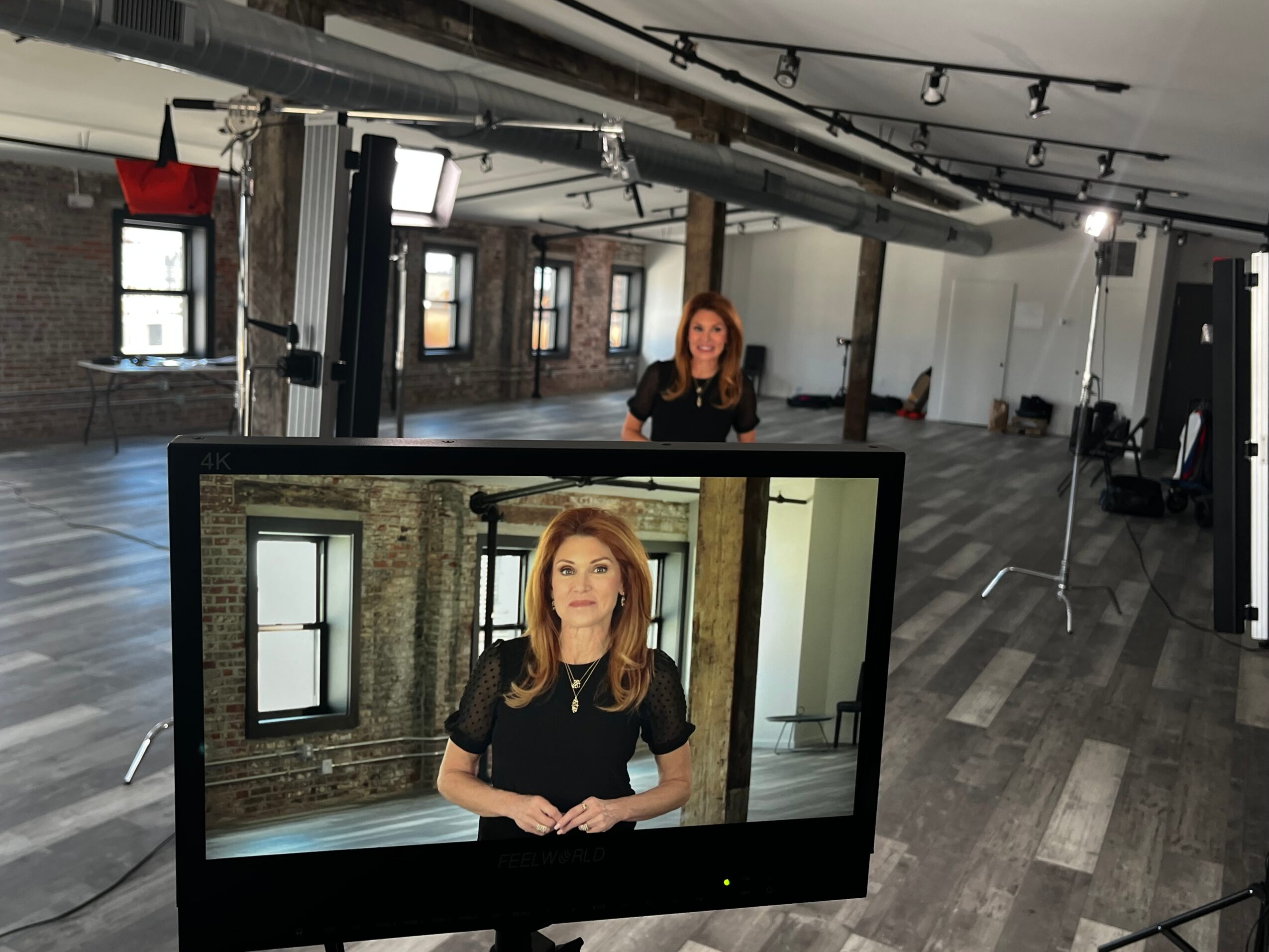 Woman standing in a studio with brick walls and large windows, seen on a monitor in the foreground.
