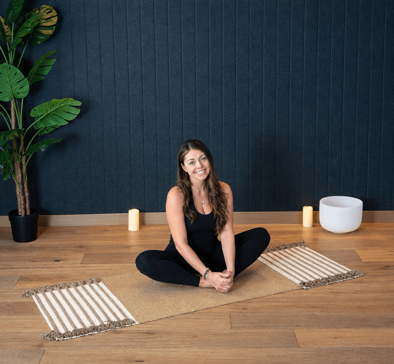 Woman sitting cross-legged on a yoga mat, smiling, in a room with a dark blue wall, wooden floor, and plants.
