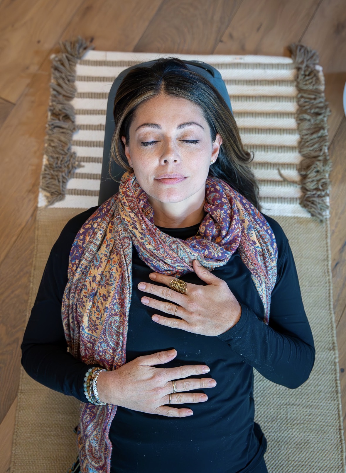 Woman lying on a striped cushion with eyes closed, wearing a colorful scarf and black top, holding her chest and stomach.