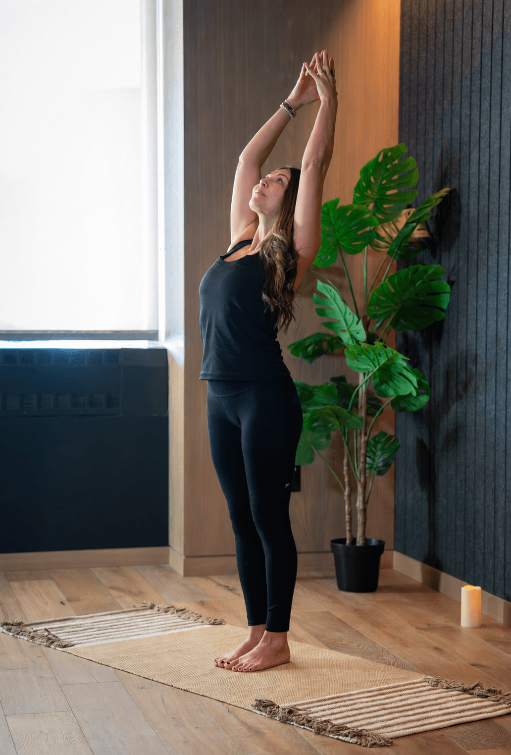 Woman standing on a yoga mat, stretching with arms raised, in a room with a large window and a potted plant.