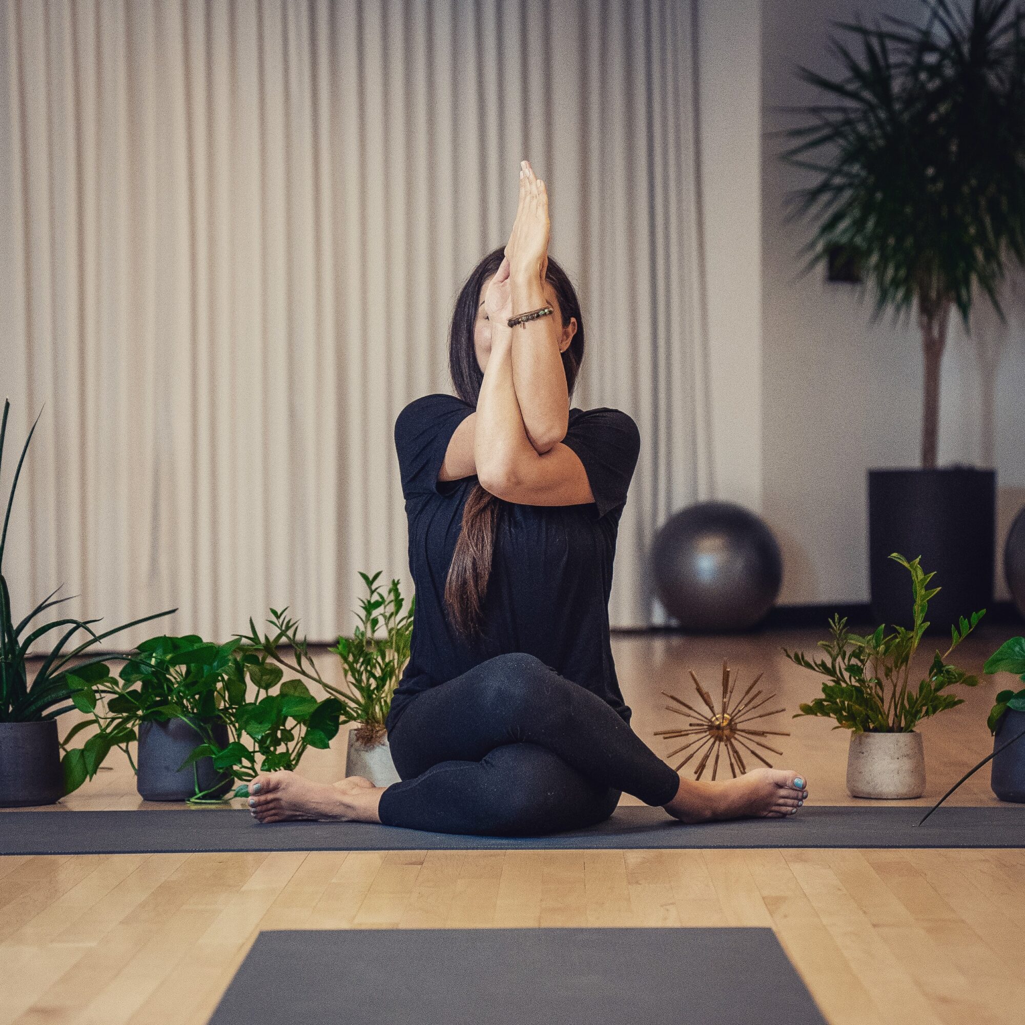 Person practicing yoga indoors, sitting cross-legged with one arm raised and the other resting on the knee, surrounded by plants.