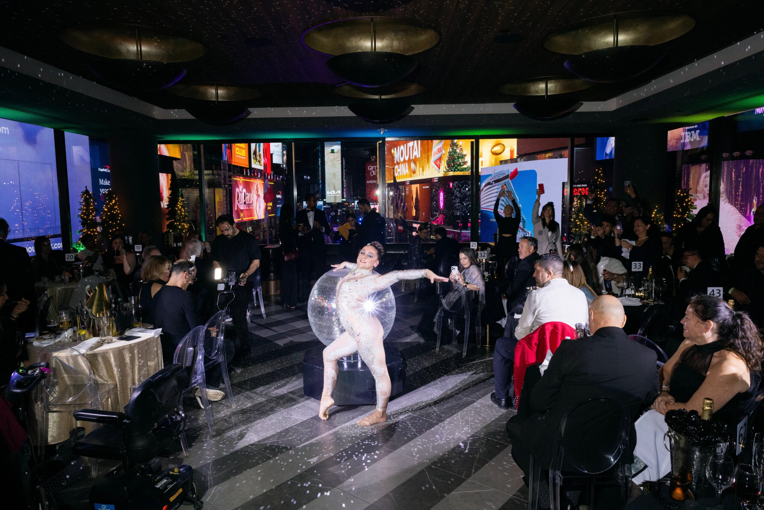 People dancing in a dimly lit venue with tables, screens, and cityscape images in the background.