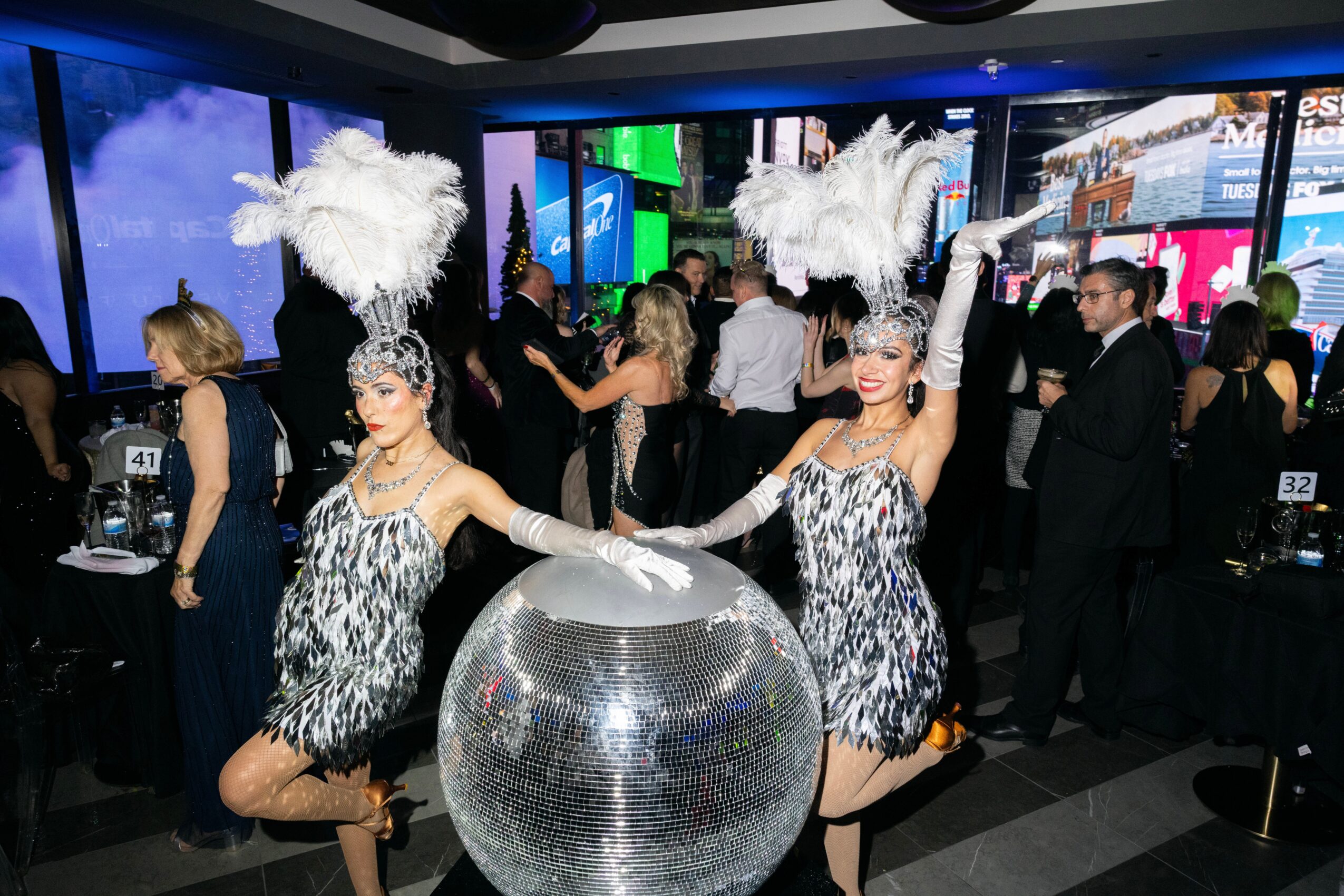 Two performers in feathered costumes pose with a large reflective sphere at an event, surrounded by guests in formal attire.