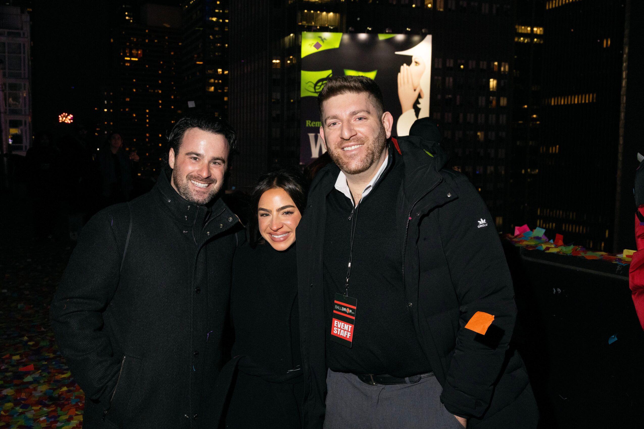 Three people smiling outdoors at night with city lights in background.
