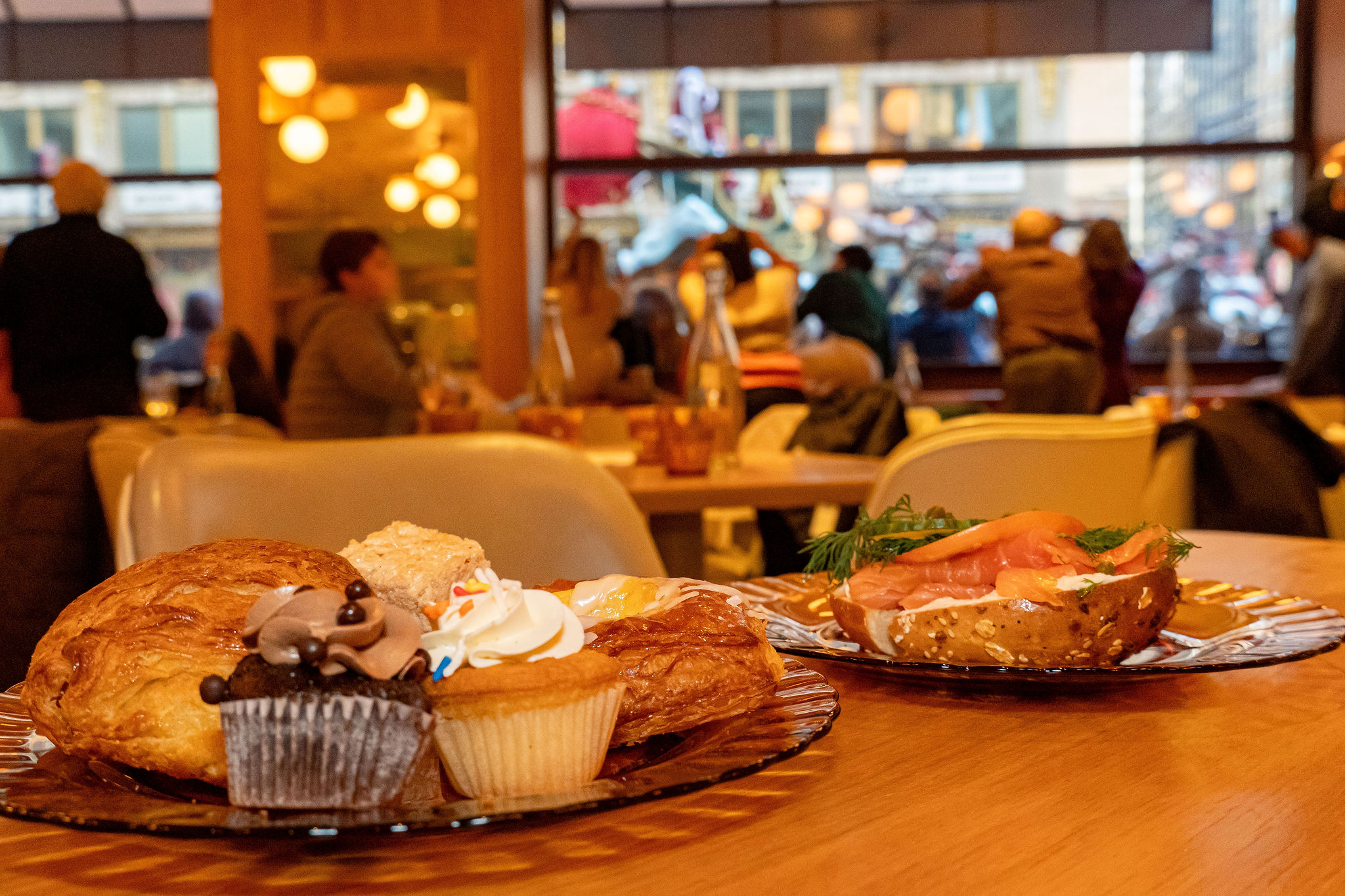 Assorted baked goods on a table in a busy cafe with people sitting and walking outside through large windows.