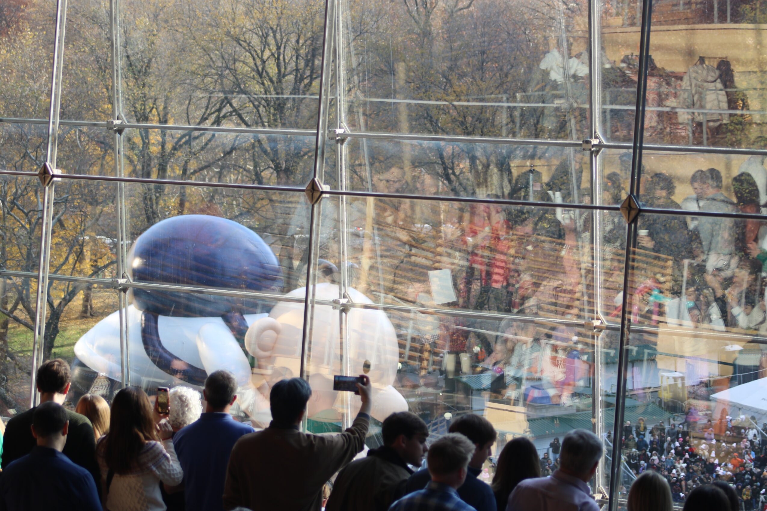 People watching a large inflatable character outside a glass building, with reflections of a crowd inside.