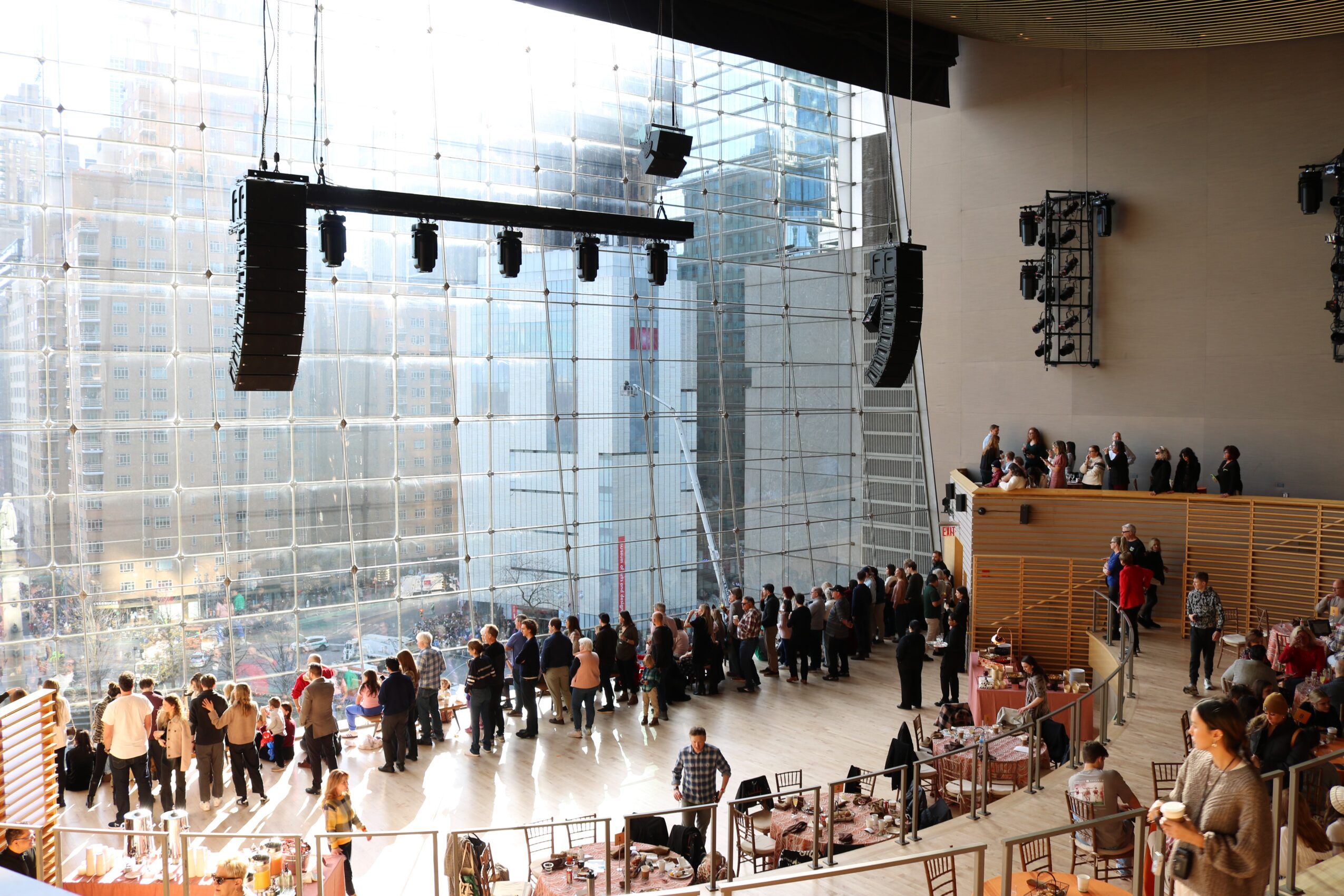 People gather inside a large modern building with glass walls and a stage with lighting equipment, some seated and others standing.