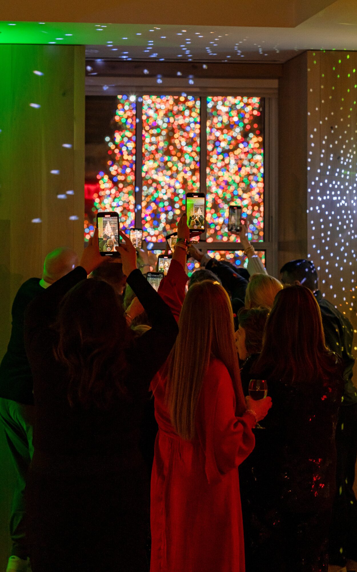 People watching a colorful light display on a window, some taking photos, in a dimly lit room.