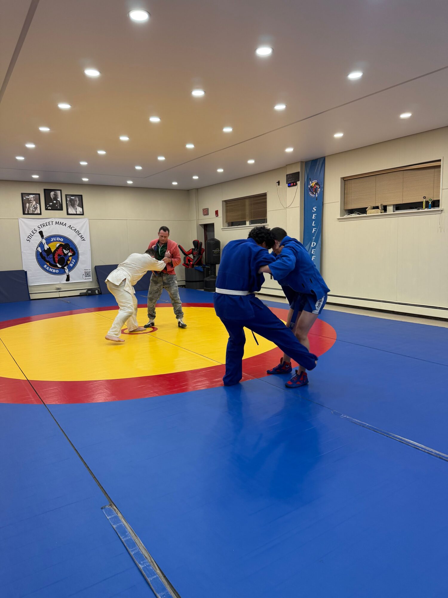 Two people practicing judo on a mat with a yellow and red circle, one in a white gi and the other in a blue gi.