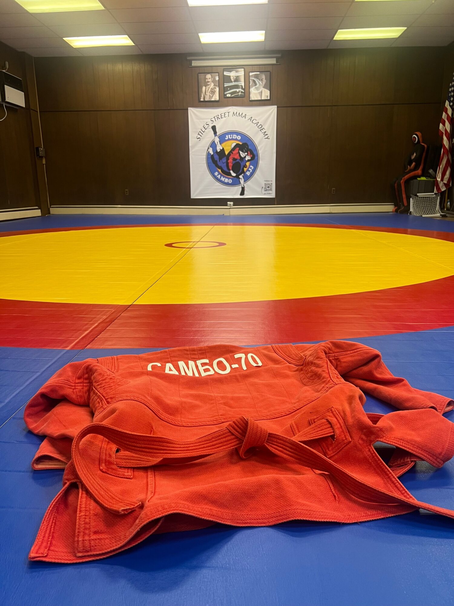 Red judo gi on blue mat with yellow and red circle, dojo wall with logo in background.