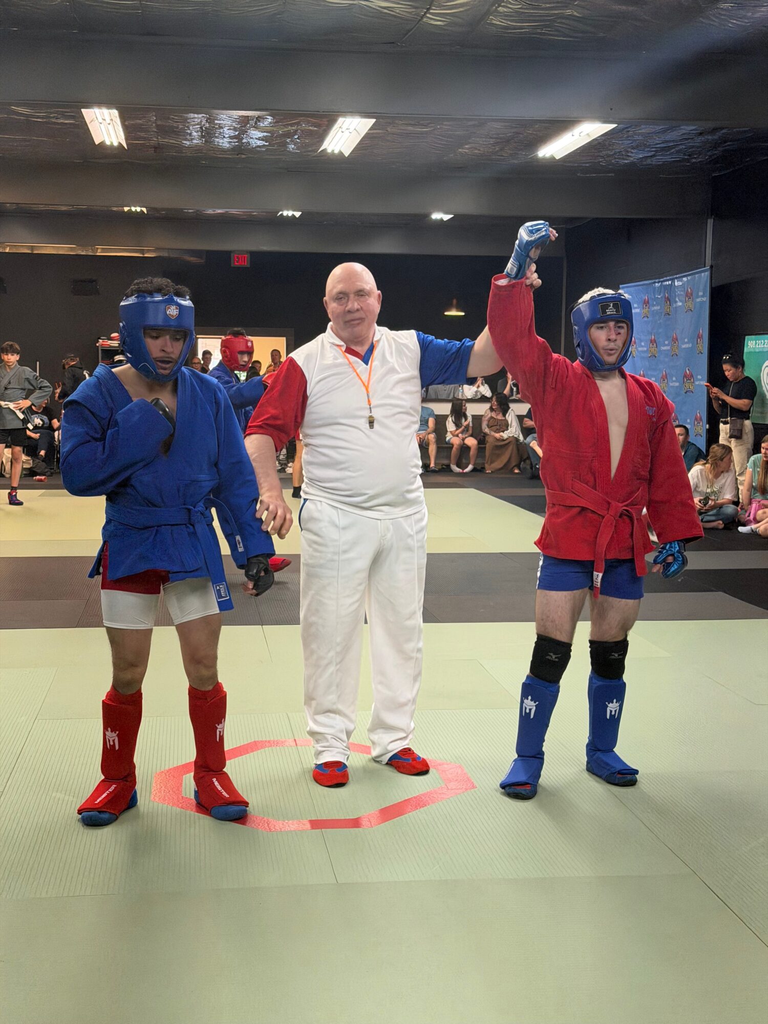 Two people in costumes stand on a green floor, one in a white outfit and the other in a blue outfit, with a crowd in the background.