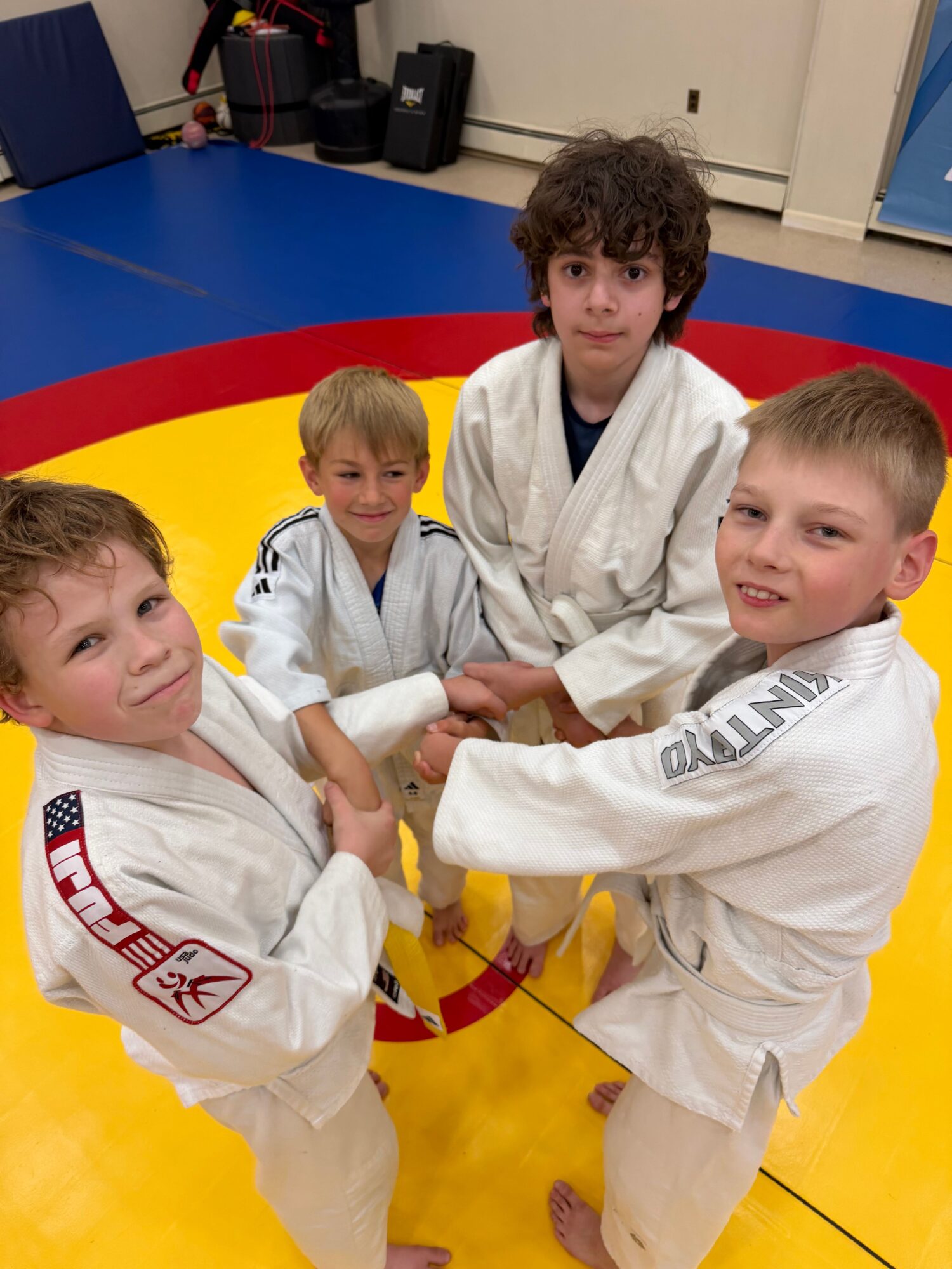 Three children practicing judo on a yellow mat, wearing white gis with belts, in a dojo setting.