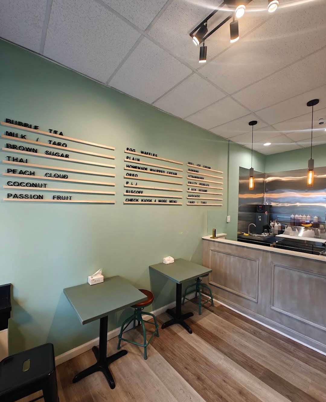 Interior of a cafe with a light green wall, two small tables, and a counter with hanging lights, wooden flooring.