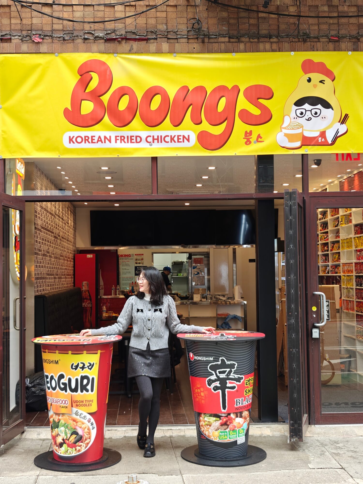 Woman standing in front of Boongs Korean fried chicken restaurant entrance, with large colorful menu containers outside.