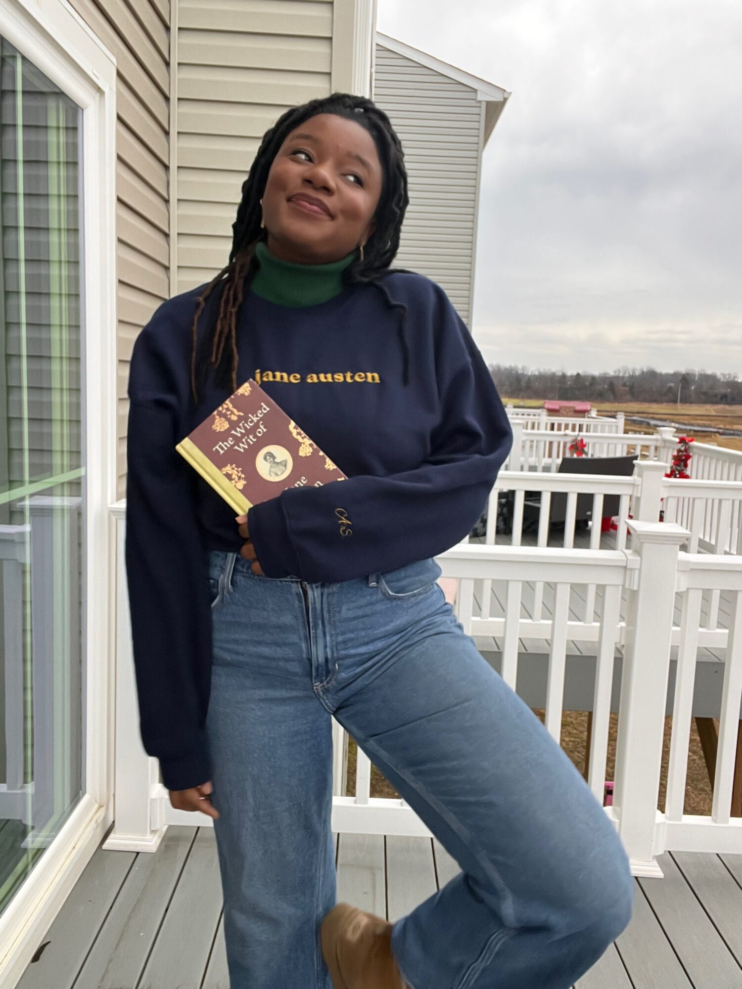 Young woman with braided hair standing on a balcony holding a book, wearing a navy sweatshirt and jeans, smiling.