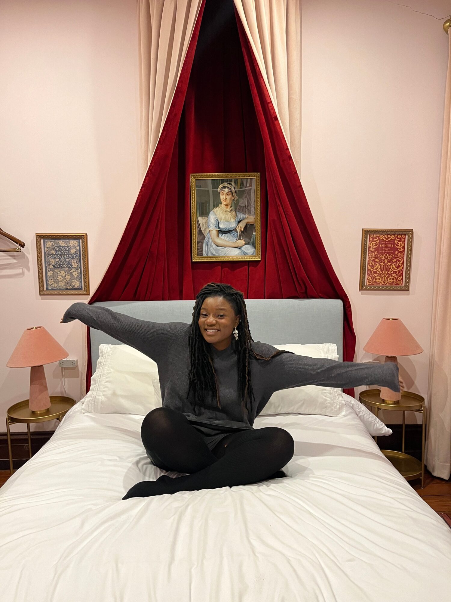Person sitting on a bed with arms outstretched, in a room with framed pictures and red curtains behind the bed.