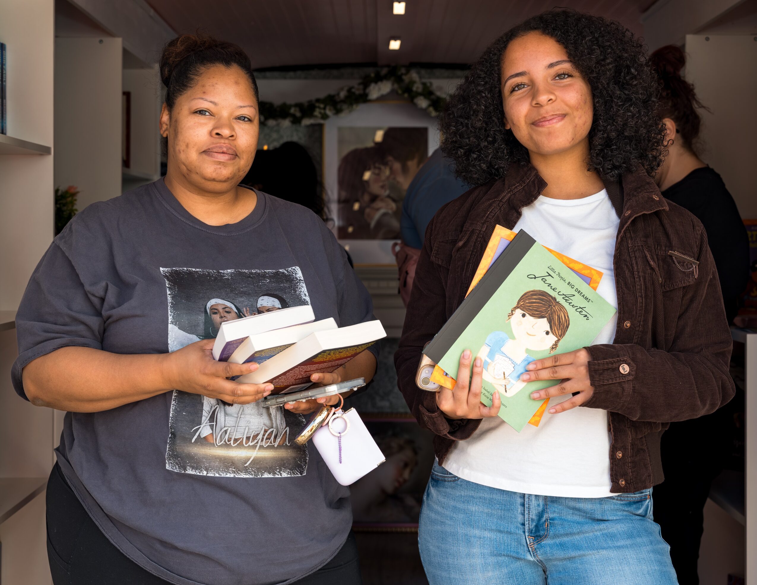 Two women holding books and smiling indoors, with people and decorations in the background.