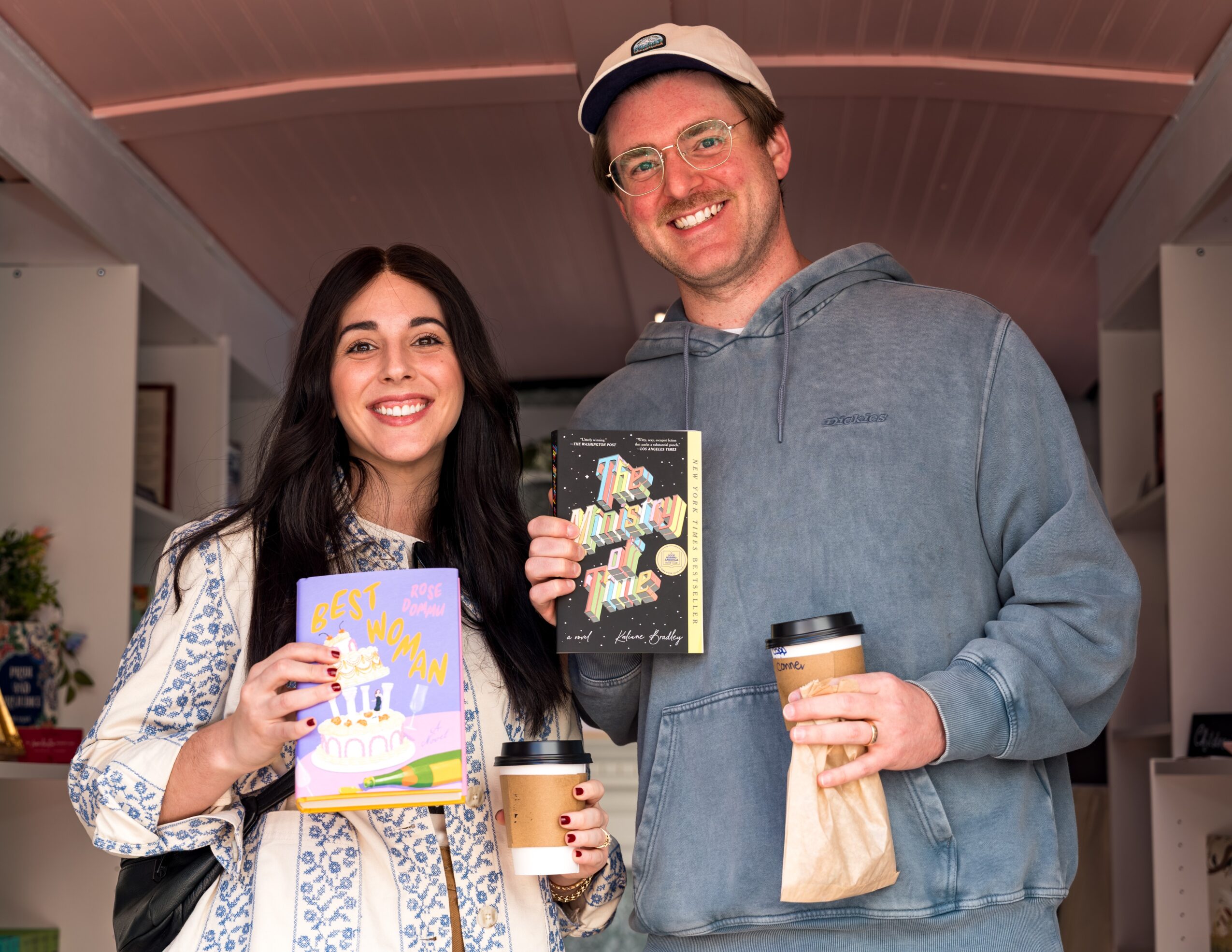 Two smiling people holding coffee cups and greeting cards inside a room with shelves and decorations.