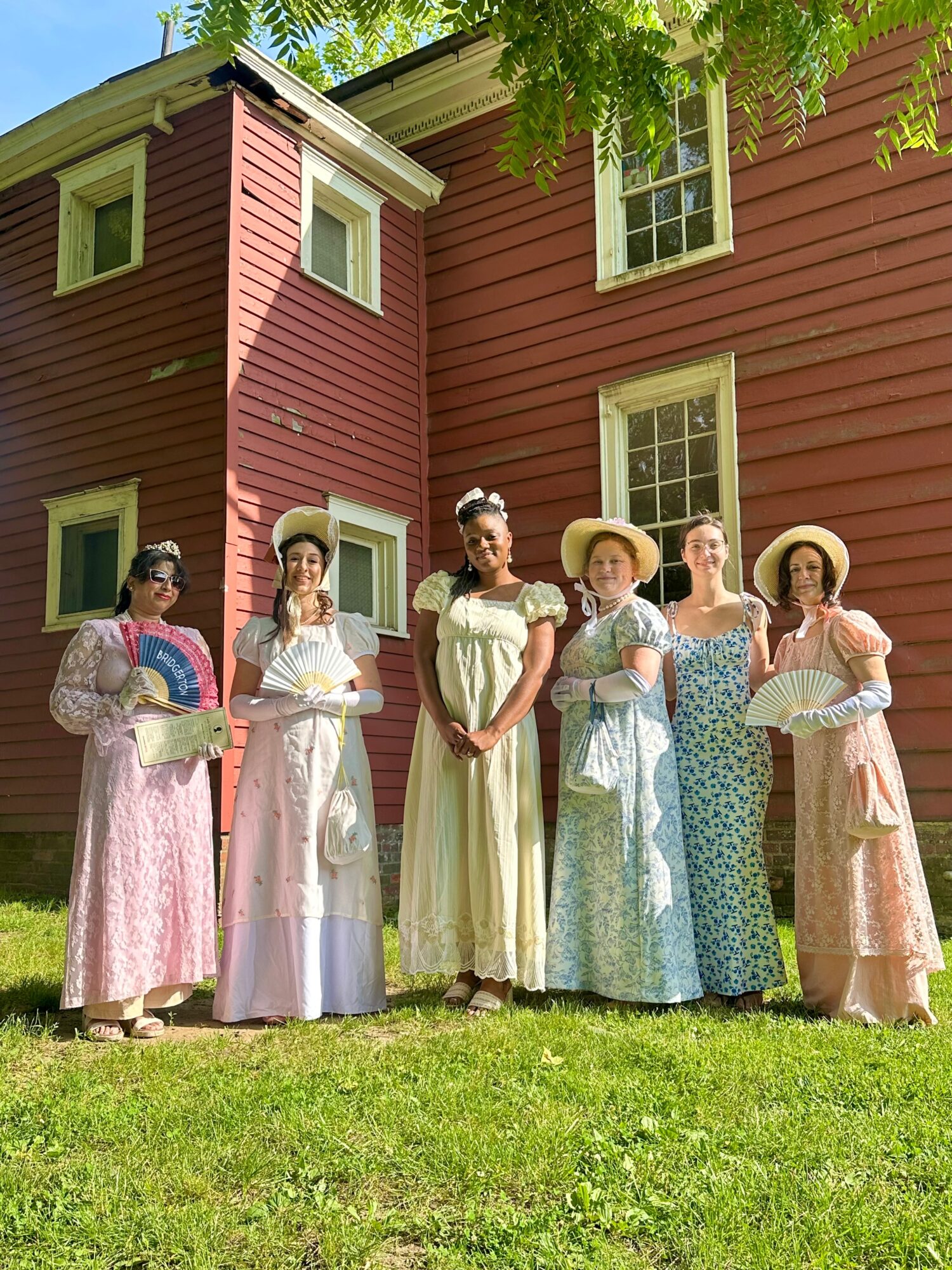 Six women in vintage dresses and hats stand on grass in front of a red wooden house with white window frames, trees overhead.