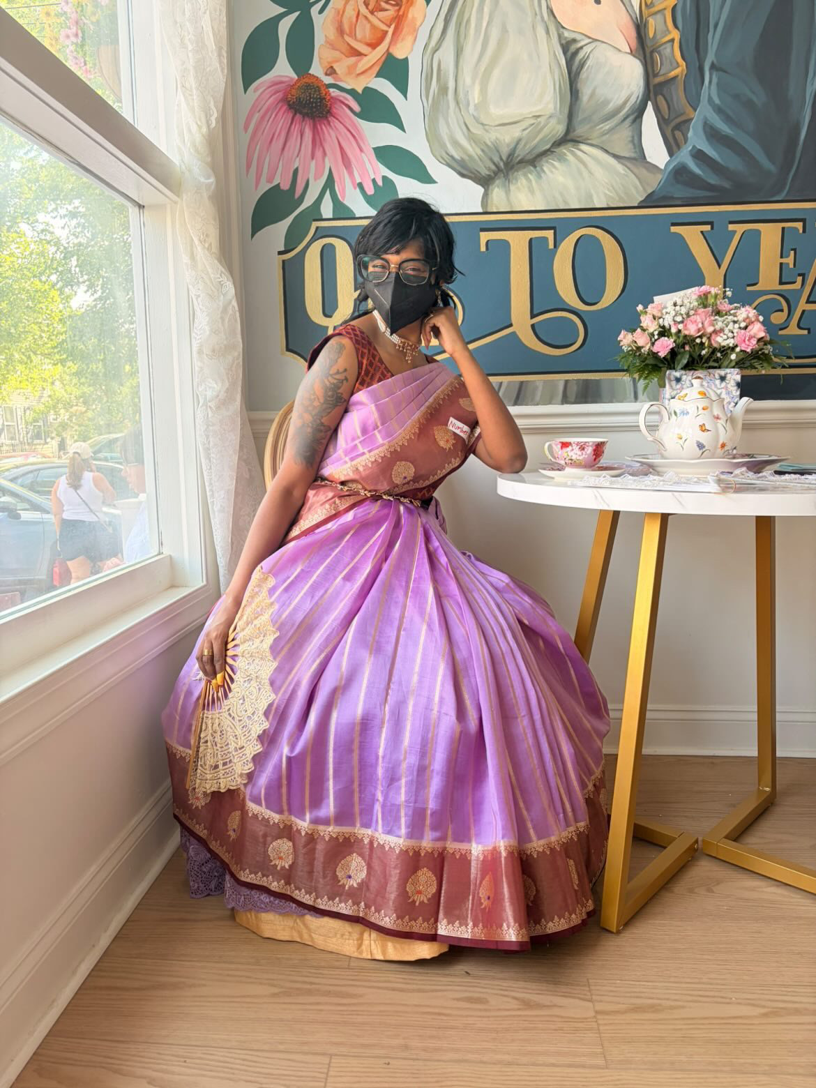 Woman in pink and purple traditional dress sitting by window with floral mural background, holding a fan, wearing a face mask.