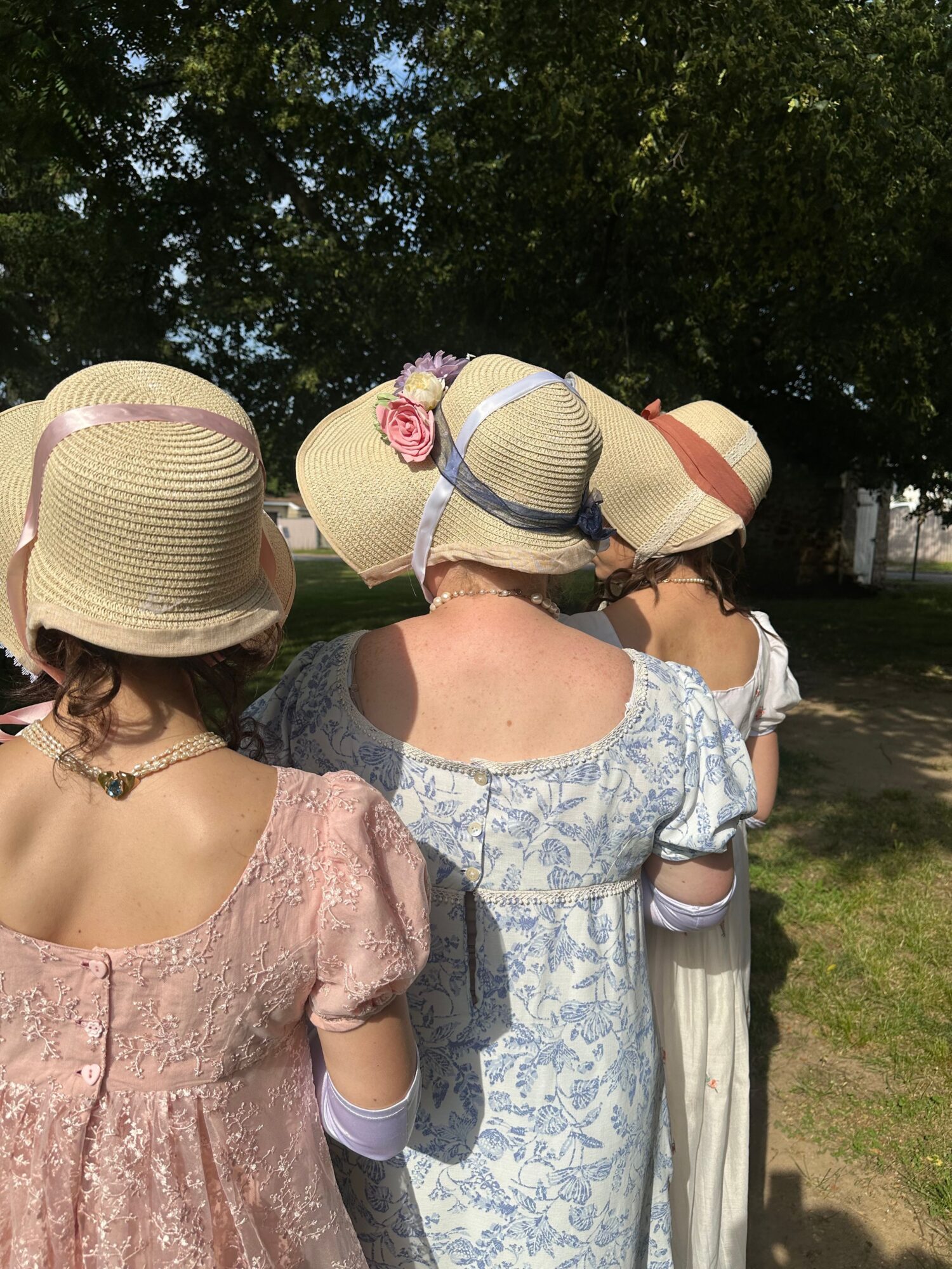 Two women in dresses and hats outdoors, one with a pink flower, facing away from the camera.