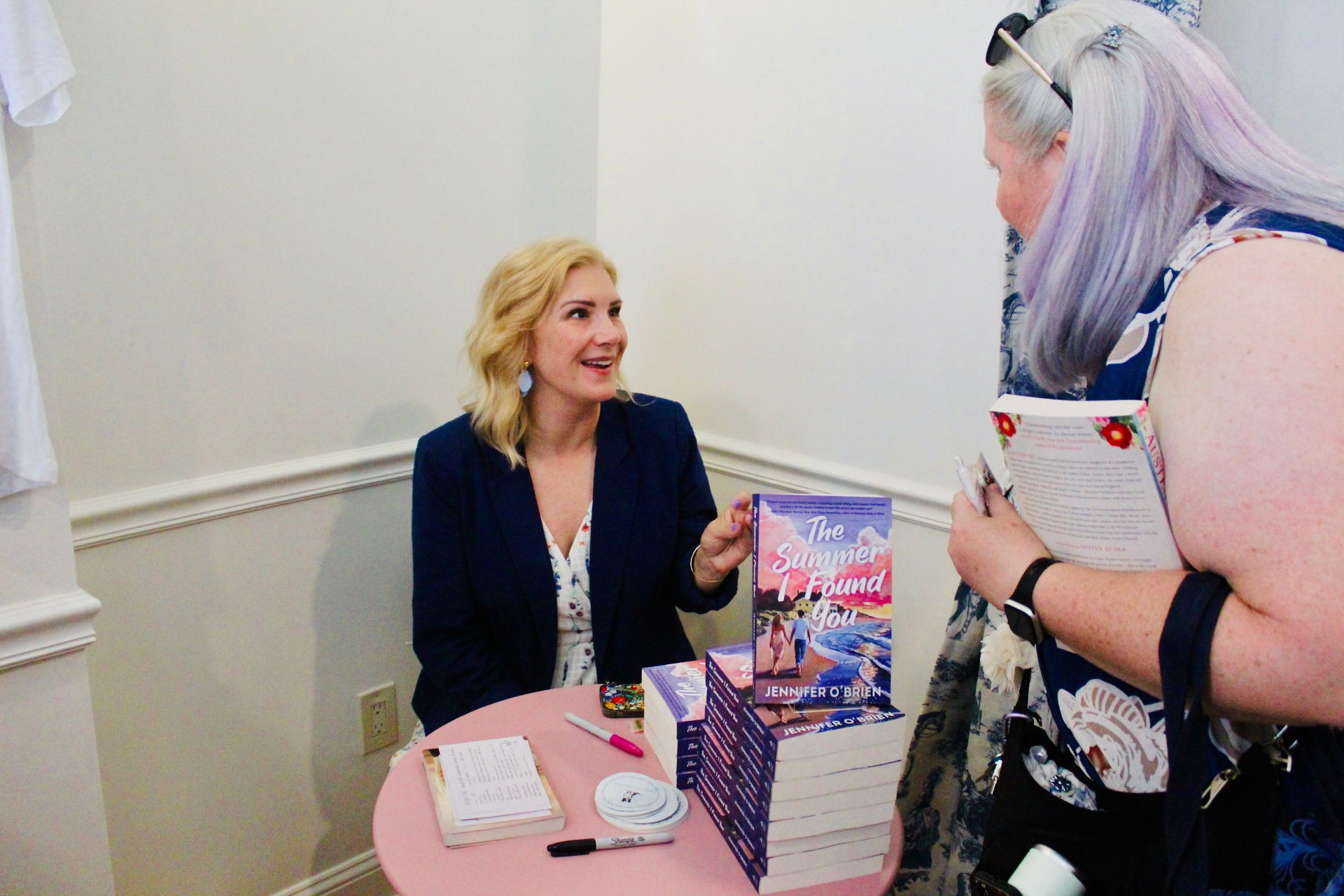 Two women talking at a table with books, one sitting and one standing, in a room with plain walls.