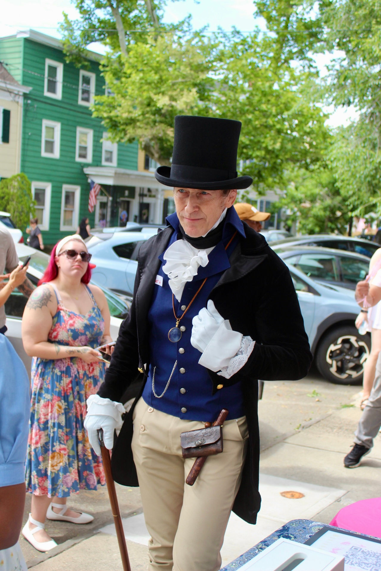 Person dressed in historical costume with top hat, blue waistcoat, and white gloves, walking outdoors on a sunny day.