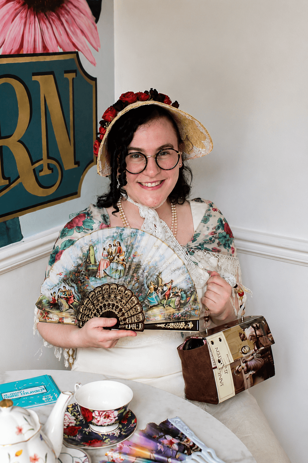 Woman wearing a hat and glasses, holding a decorative fan, sitting at a table with teapot and cups, smiling.