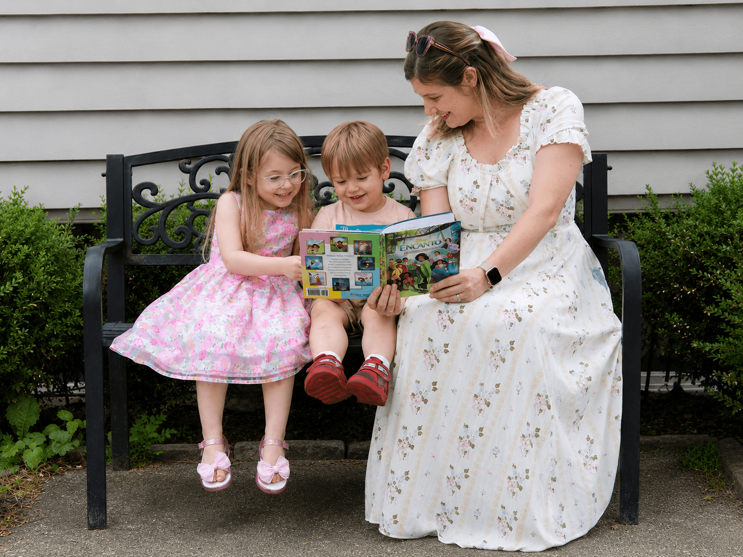 Woman reading a book to two children sitting on a bench outside, with a gray house in the background.