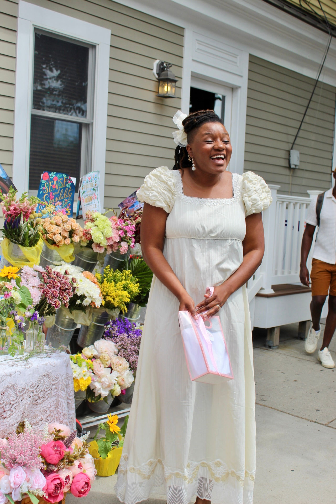 Woman in white dress smiling outdoors near colorful flower arrangements, holding a pink cloth, in front of a house with beige siding.