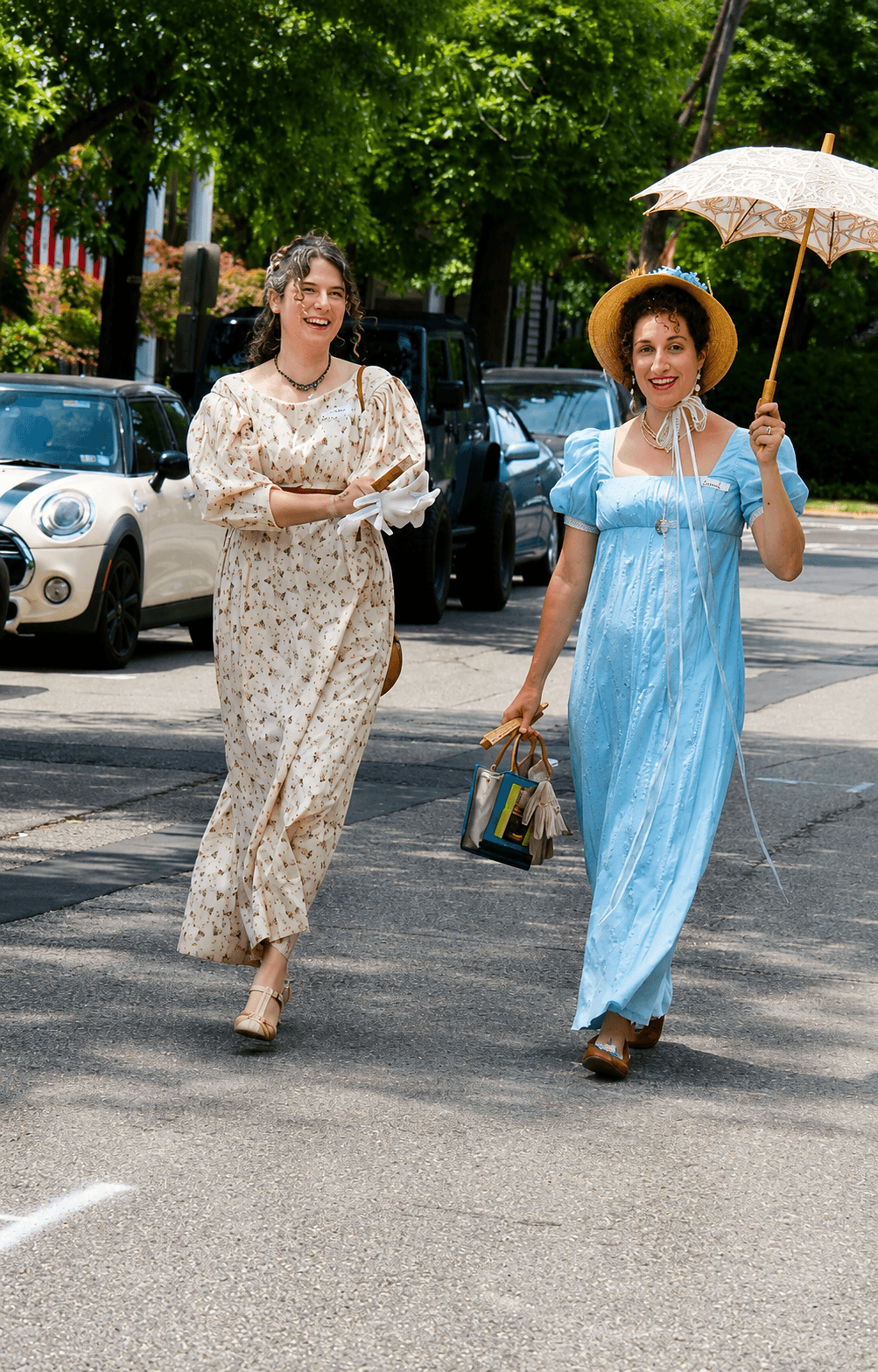 Two women walking on a street, one holding an umbrella and a bag, the other smiling with arms crossed, trees and cars in background.