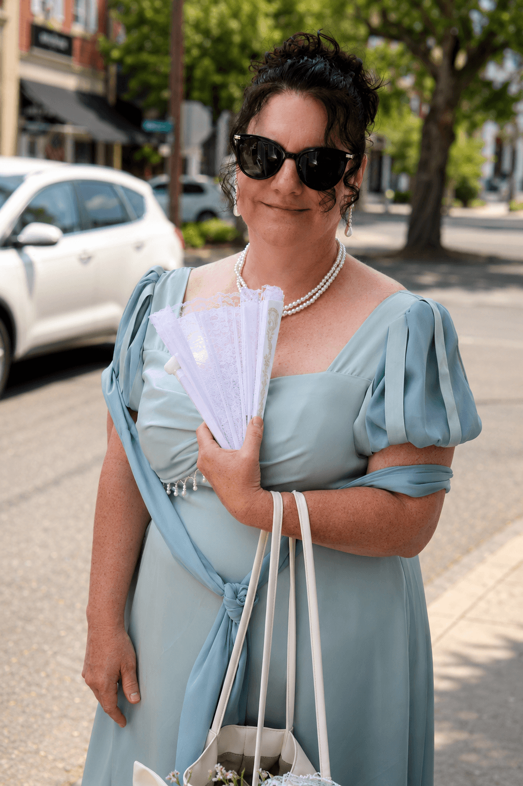 Woman in sunglasses and a light blue dress holding a fan and shopping bags on a city sidewalk.