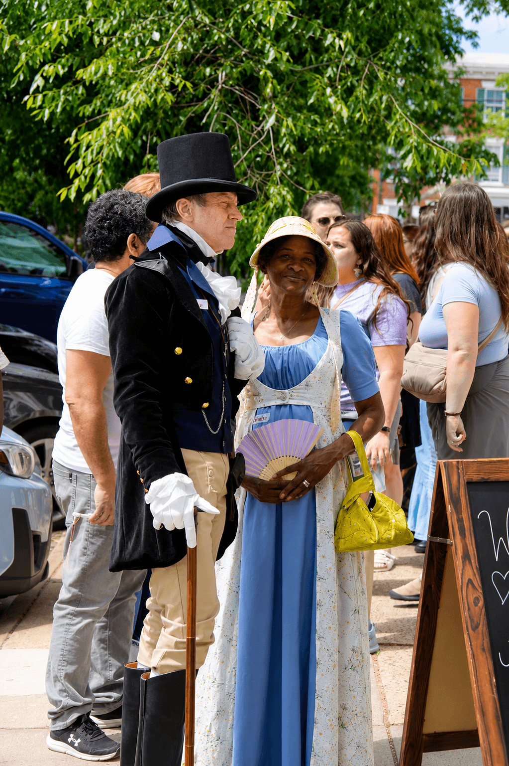 Group of people outdoors, some dressed in historical costumes, standing near a chalkboard sign, with trees and buildings in background.