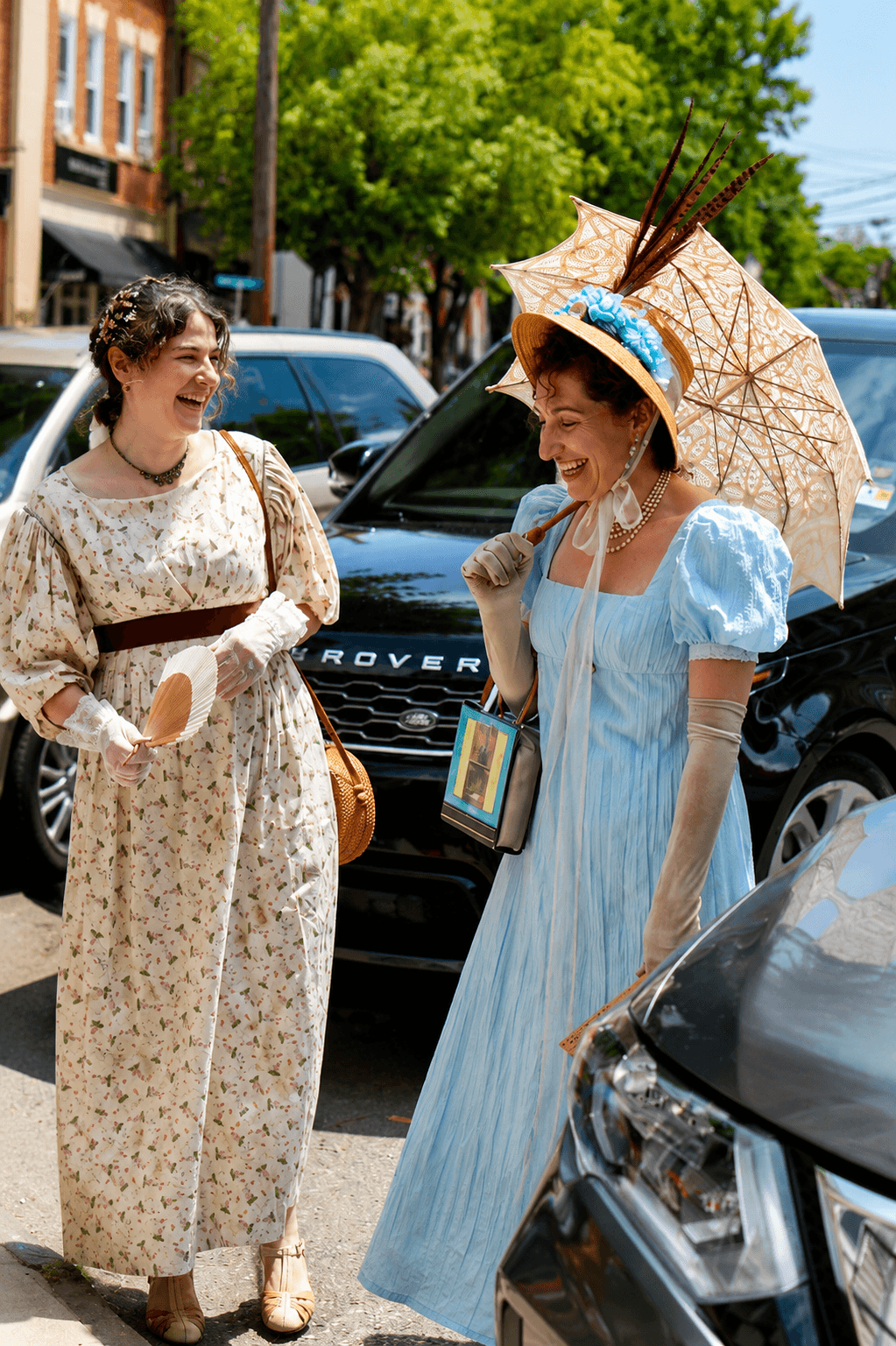 Two women in vintage dresses smiling and talking outdoors, one holding a parasol and wearing a hat, cars and trees in background.