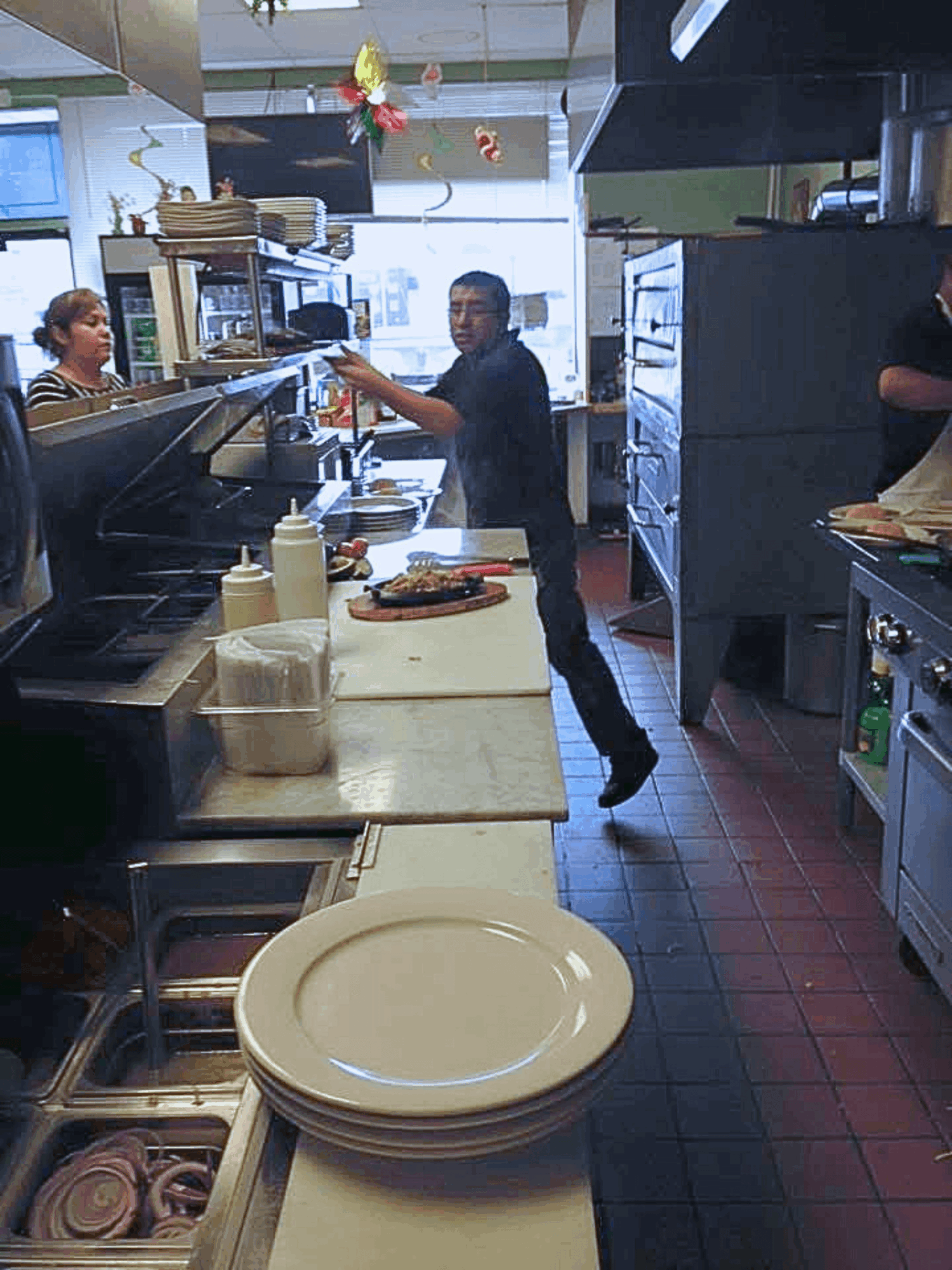 Person behind the counter serving a customer in a restaurant kitchen, with plates and kitchen equipment visible.