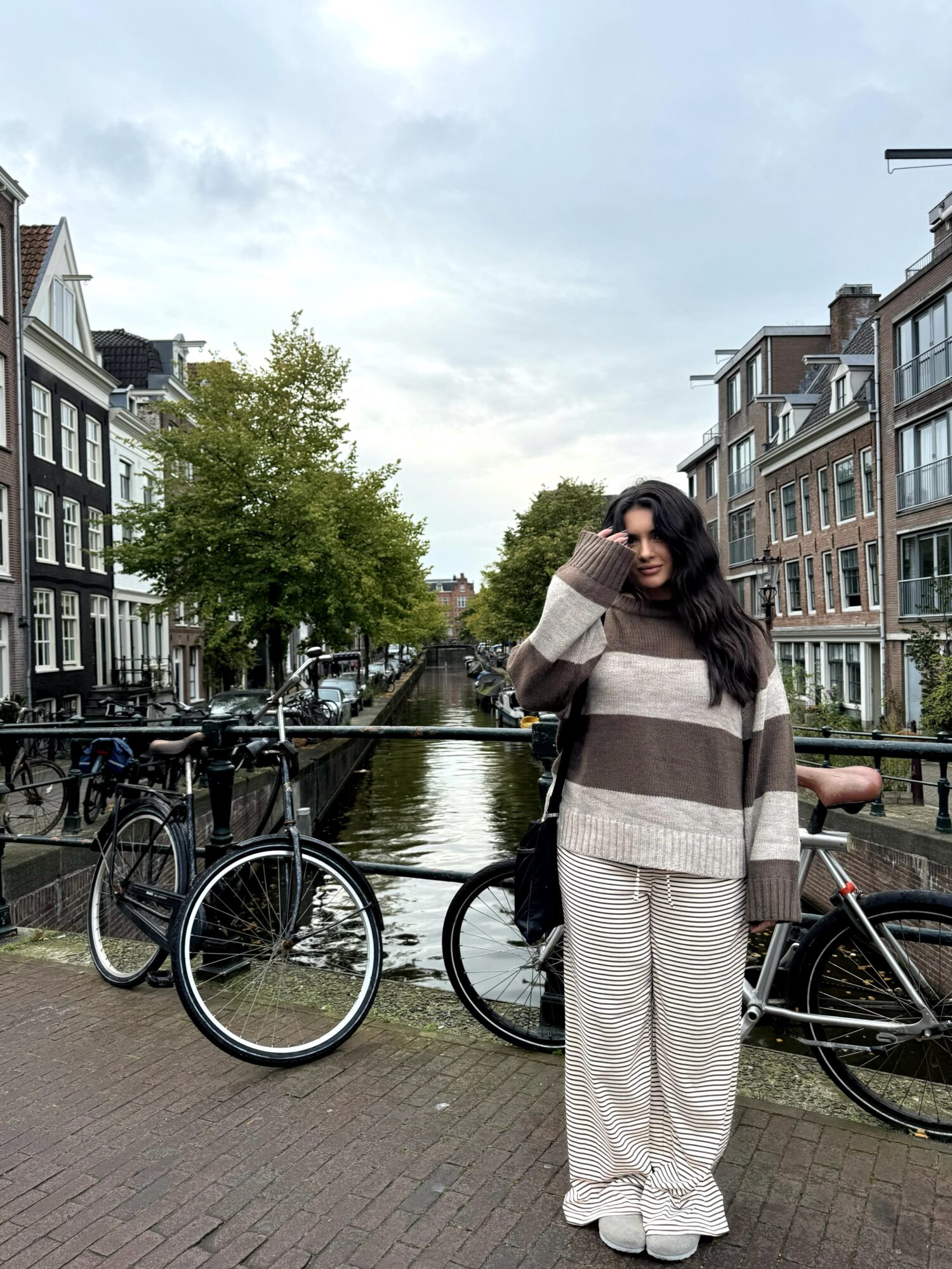 Woman with long dark hair standing by canal with bicycles, in urban area with buildings and trees, overcast sky