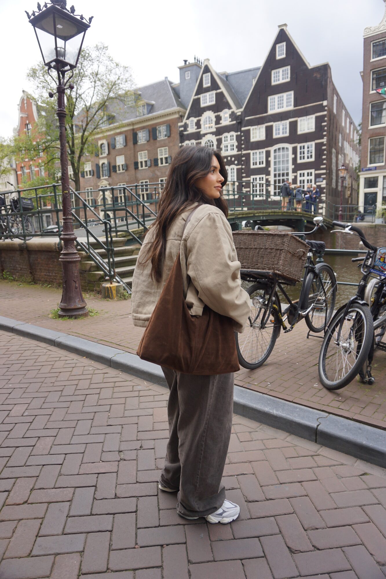Woman with long hair wearing a beige jacket and gray pants standing on a brick sidewalk near bicycles and canal, historic buildings in background.