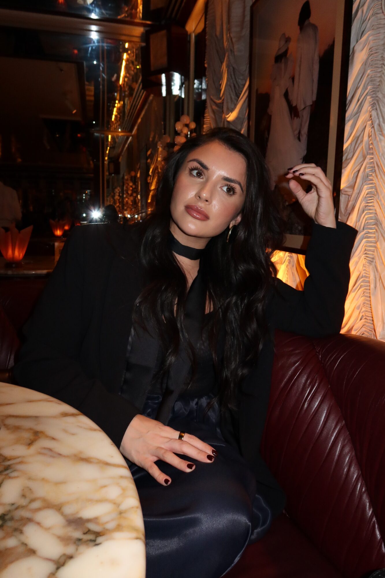 Woman with long dark hair and makeup sitting in a dimly lit restaurant or bar, wearing a black outfit and choker necklace.