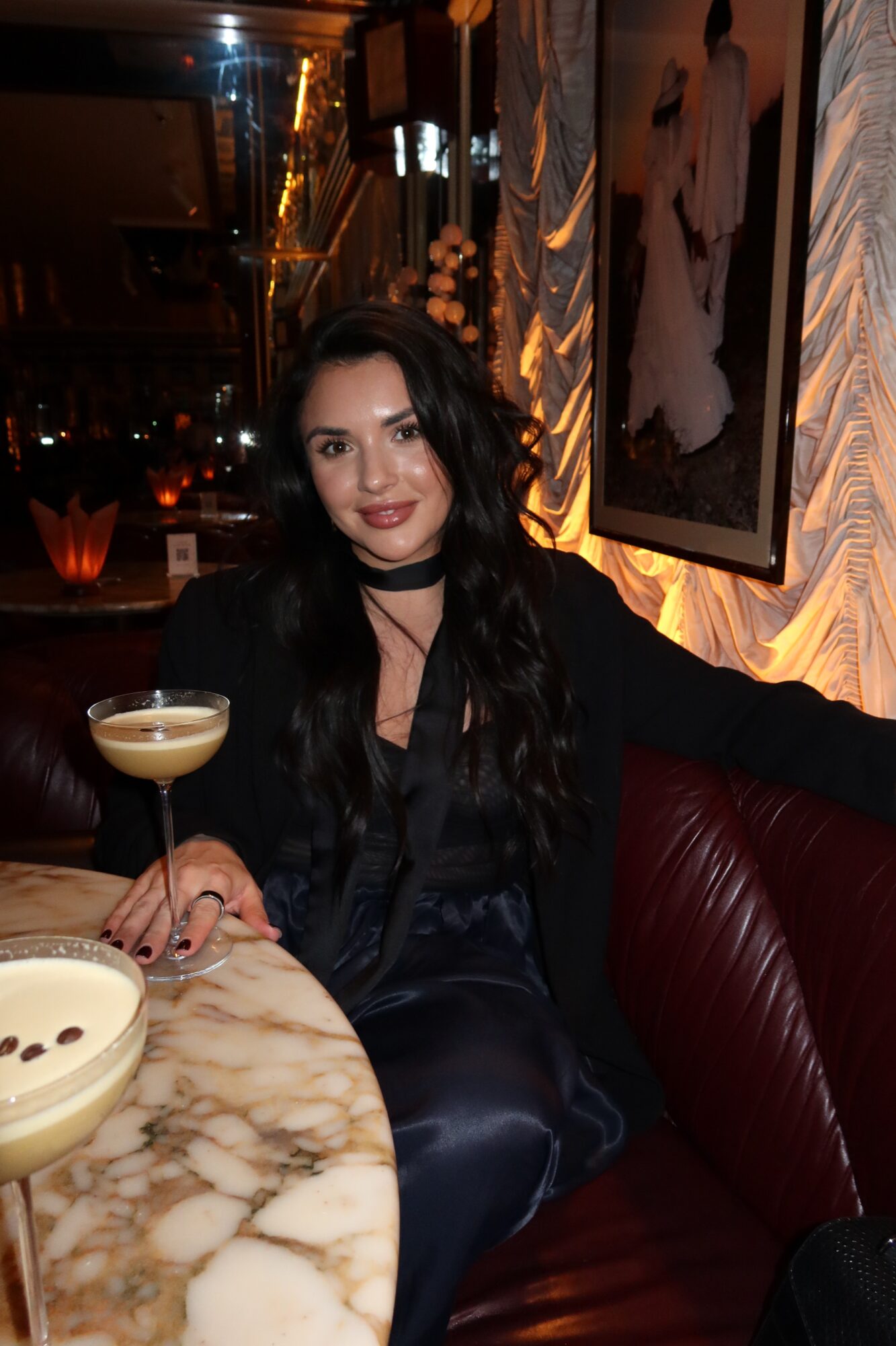 Woman with long dark hair and dark clothing sitting in a dimly lit restaurant, holding a cocktail glass, smiling.