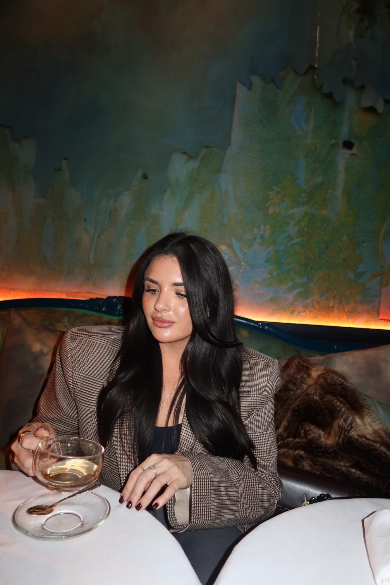 Woman with long dark hair sitting at a table, holding a glass, in front of a colorful abstract wall.