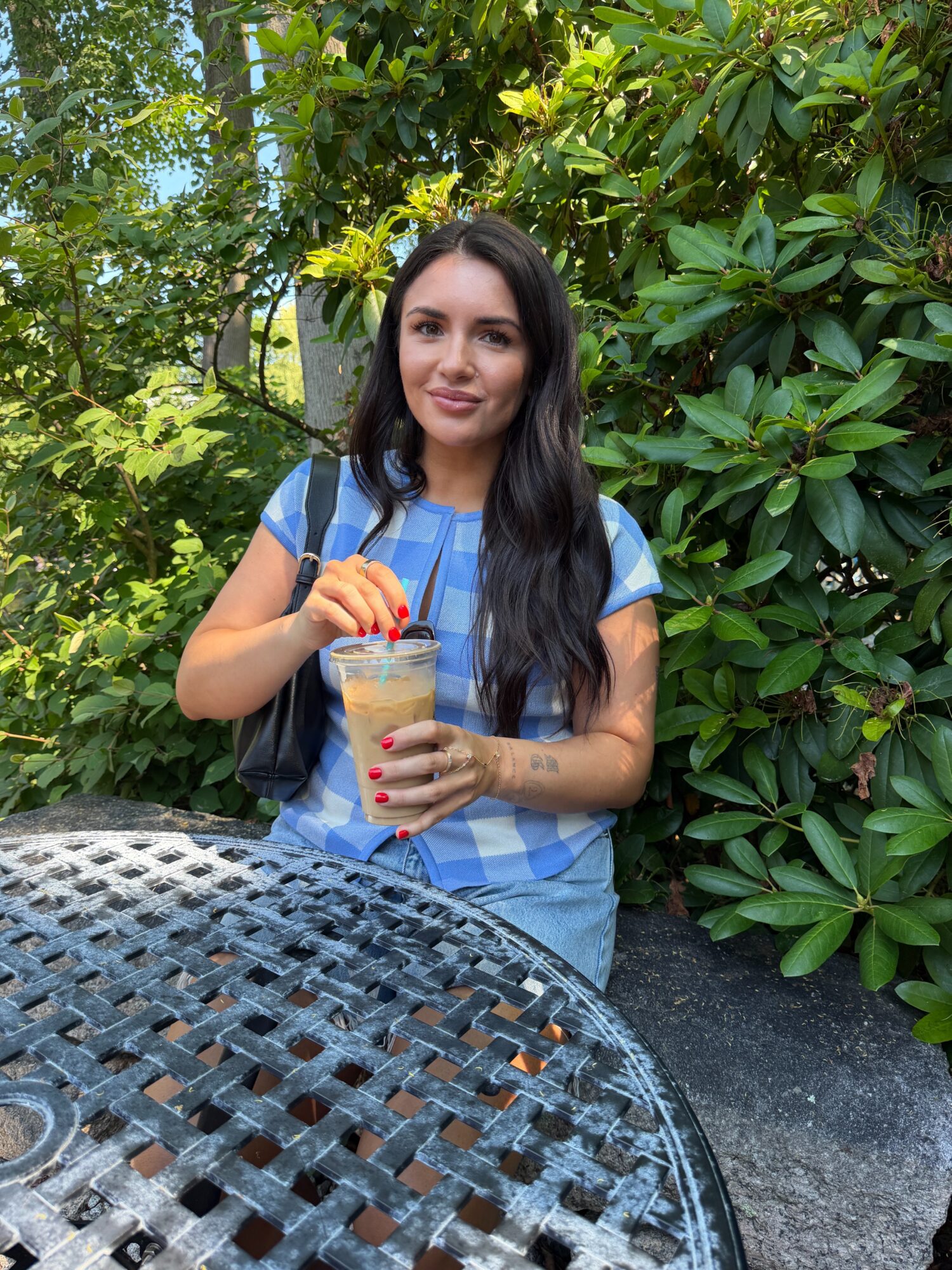 Young woman with long dark hair holding a drink with a straw outdoors, surrounded by green foliage.