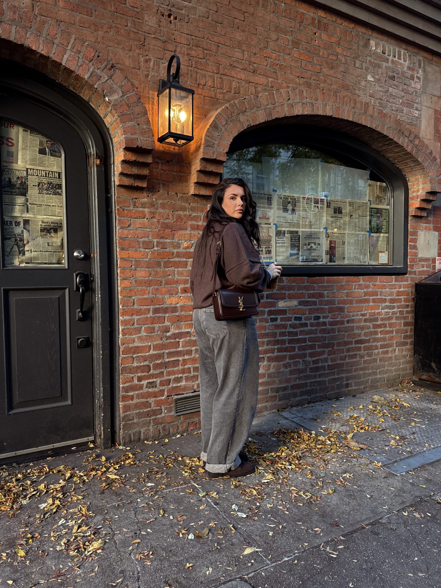 Woman standing outside brick building with window and lantern, holding a phone, leaves on ground.