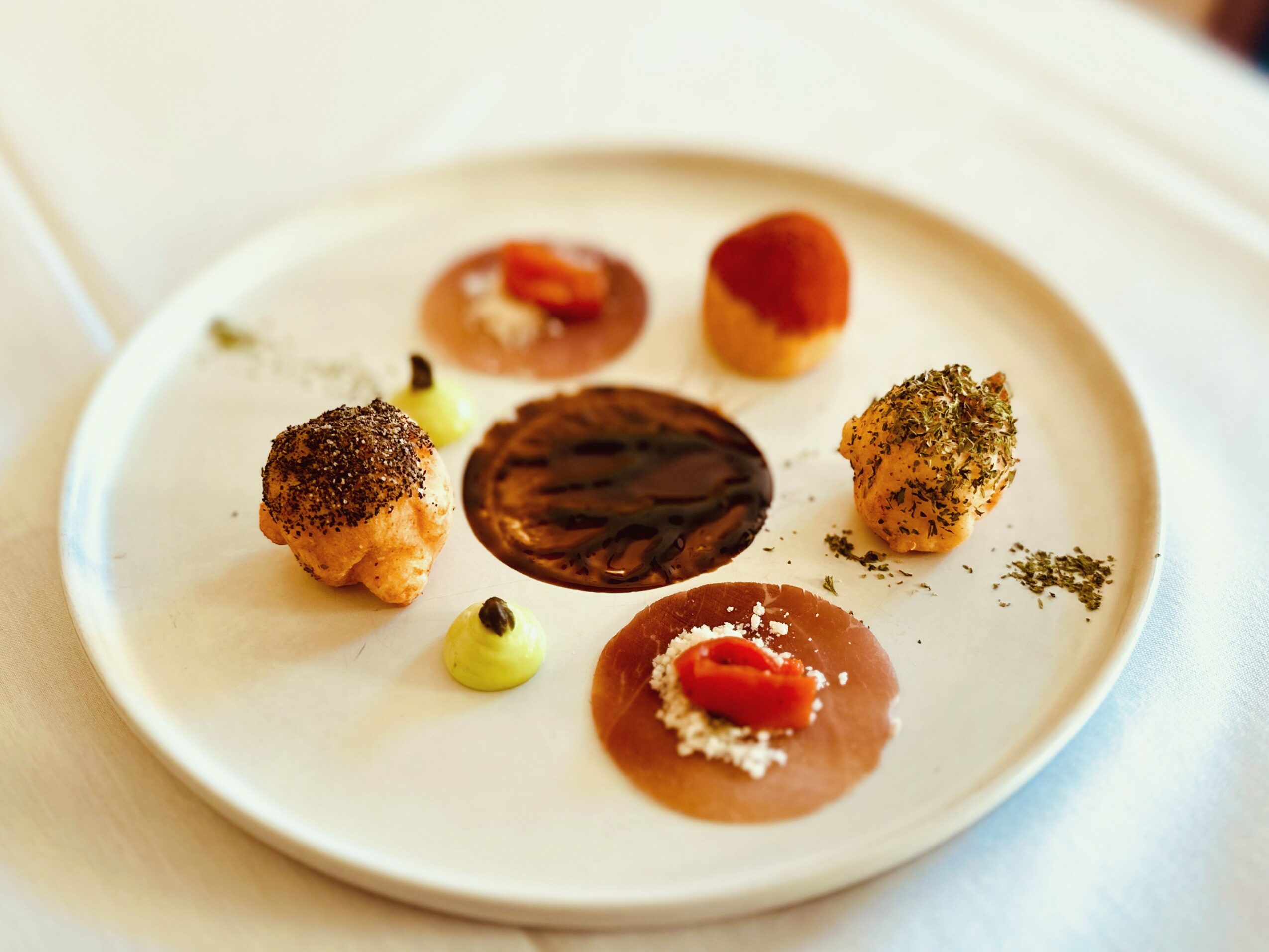 Assorted small food items on a white plate, including a dark chocolate piece, a round pastry, and various garnishes, on a light background.