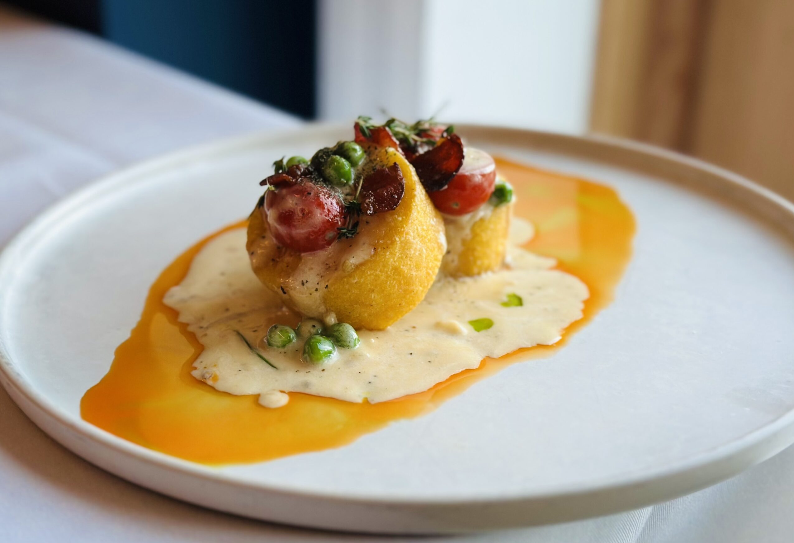 Two small, round, yellow appetizers topped with berries and herbs on a white plate with sauce, garnished with green peas.