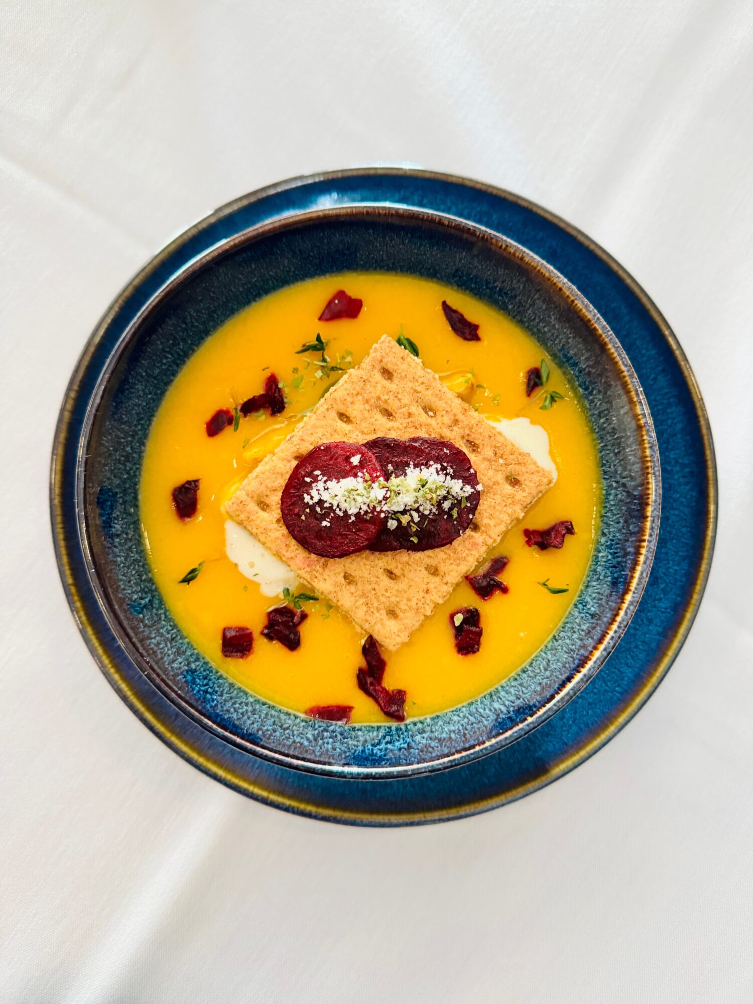 A bowl with yellow sauce, topped with a rectangular cracker, a dollop of red jam, and garnished with herbs and dried petals.