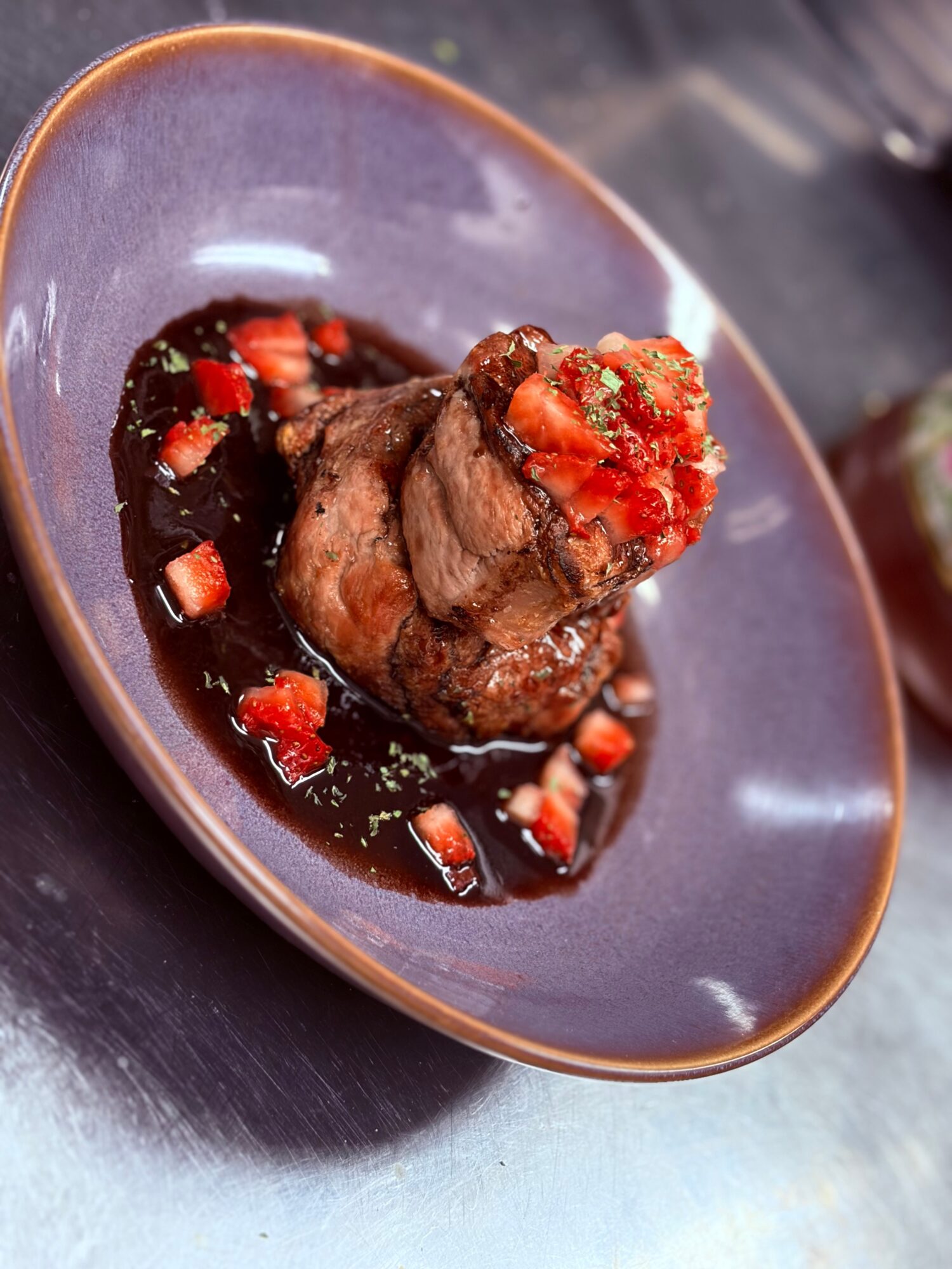 Cooked meat with sauce and chopped vegetables on a dark plate, viewed from the side.