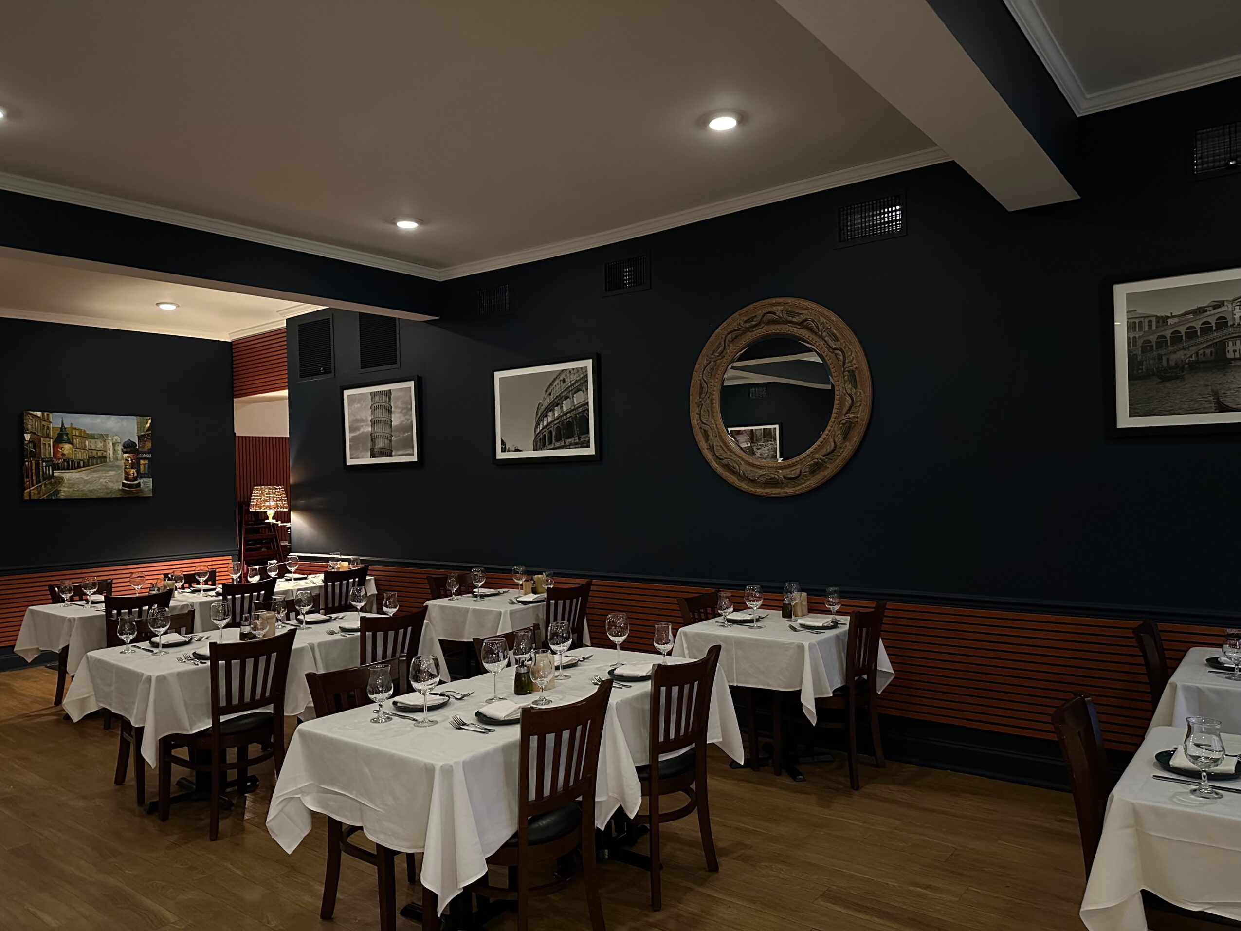 Dining room with tables, chairs, and framed pictures on a dark wall, illuminated by ceiling lights.