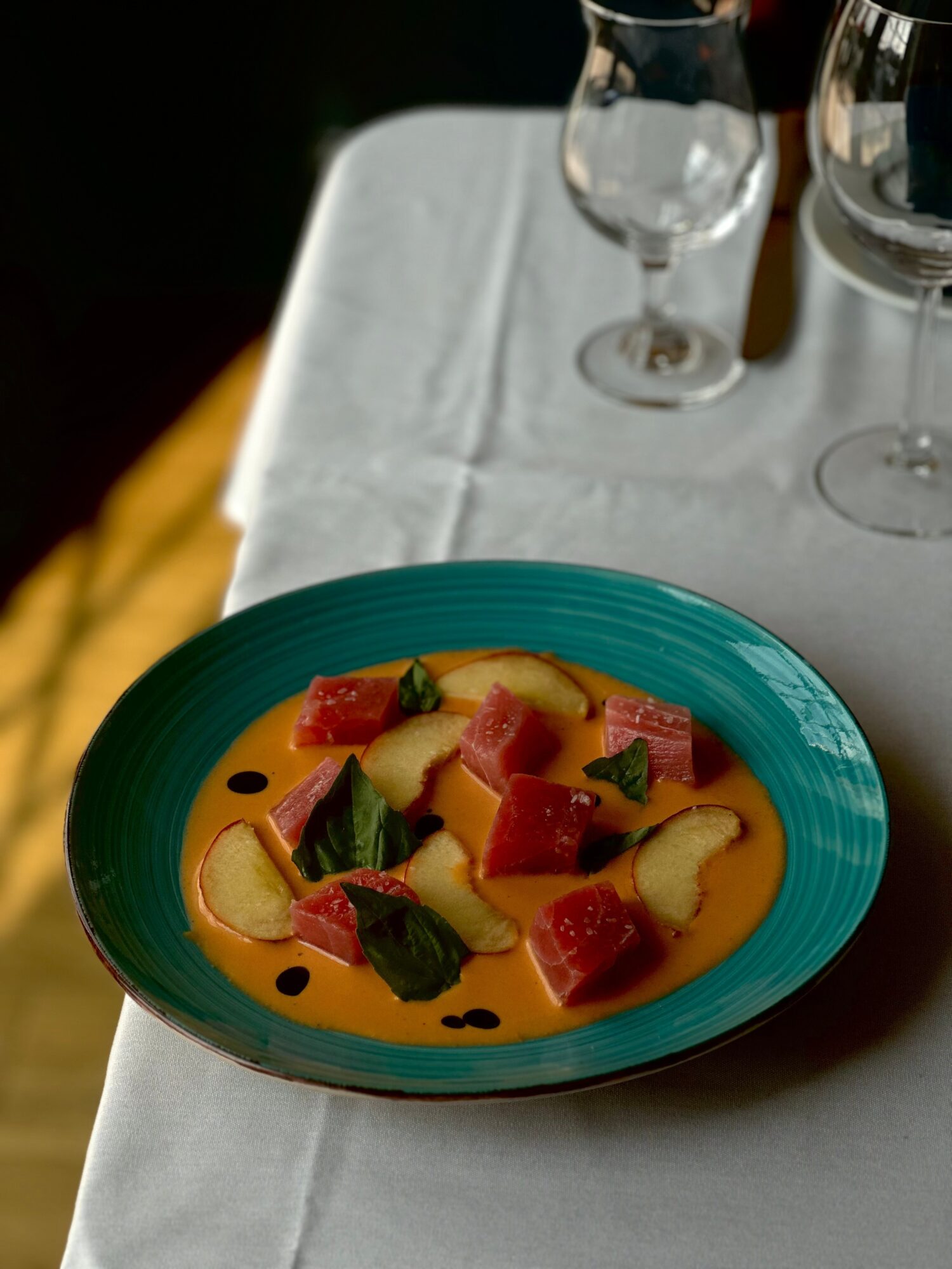 Colorful dish with chunks of fruit and sauce on a teal plate, on a white tablecloth with glasses in background.
